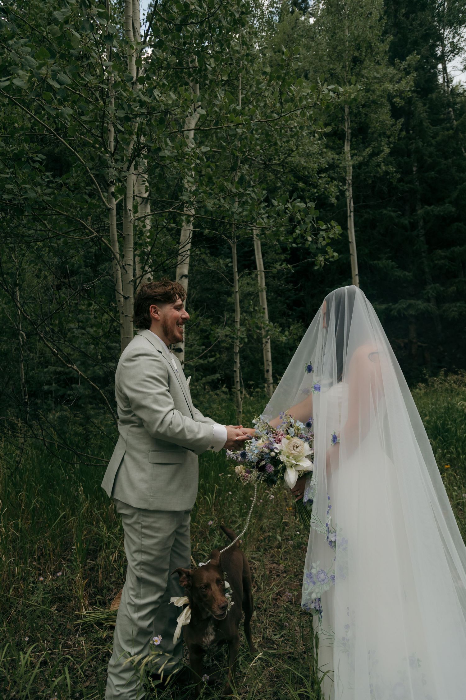 Groom in beige suit holds bouquet during forest elopement first look with bride in floral veil.