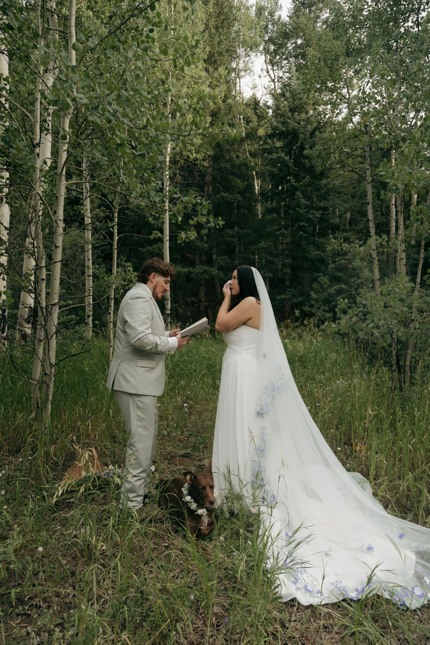 Groom reads heartfelt vows to emotional bride in white floral gown during forest elopement ceremony.