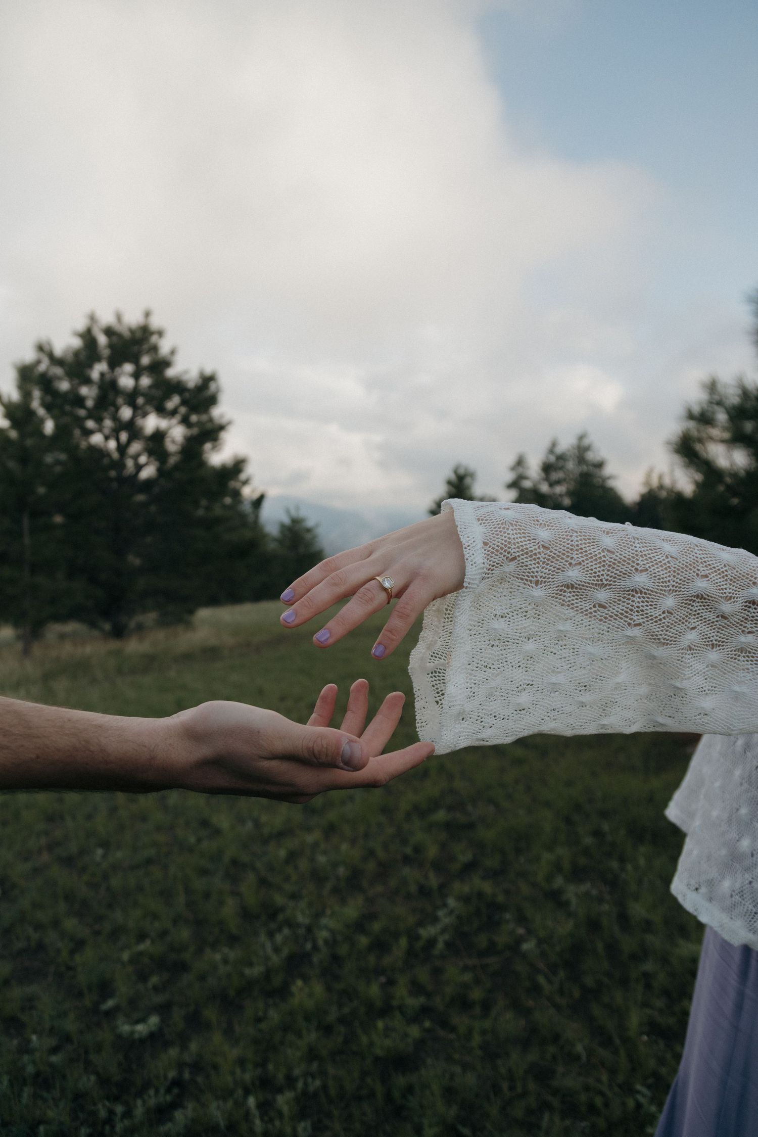Close-up of engaged couple holding hands showing diamond ring in lace sleeve outdoors.
