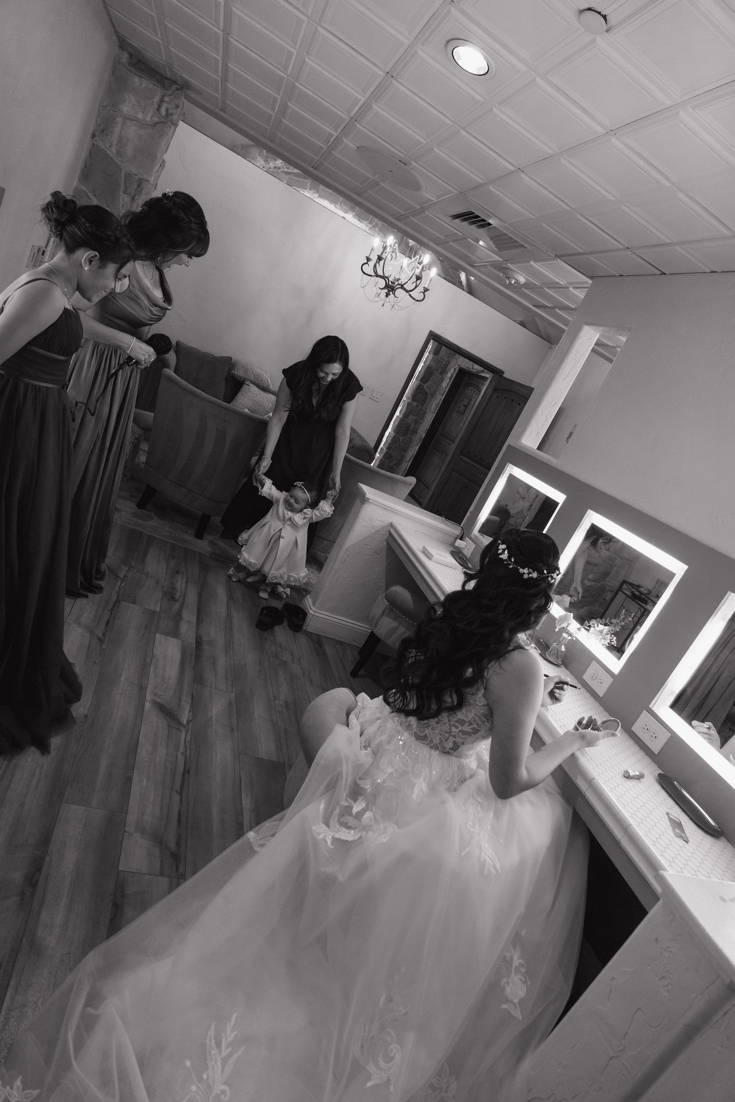Bride in white gown sits at vanity mirror as bridesmaids prepare in dressing room, black and white photo.