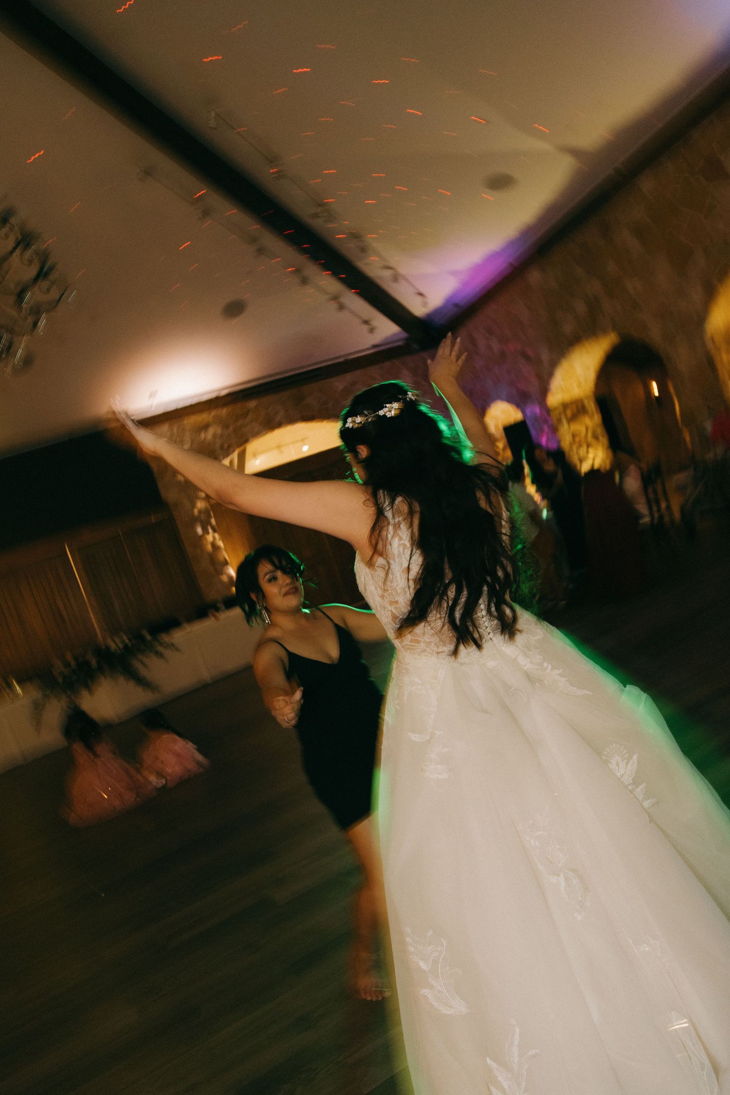 A bride in a white ball gown dances with arms raised under colorful lights at her wedding reception.