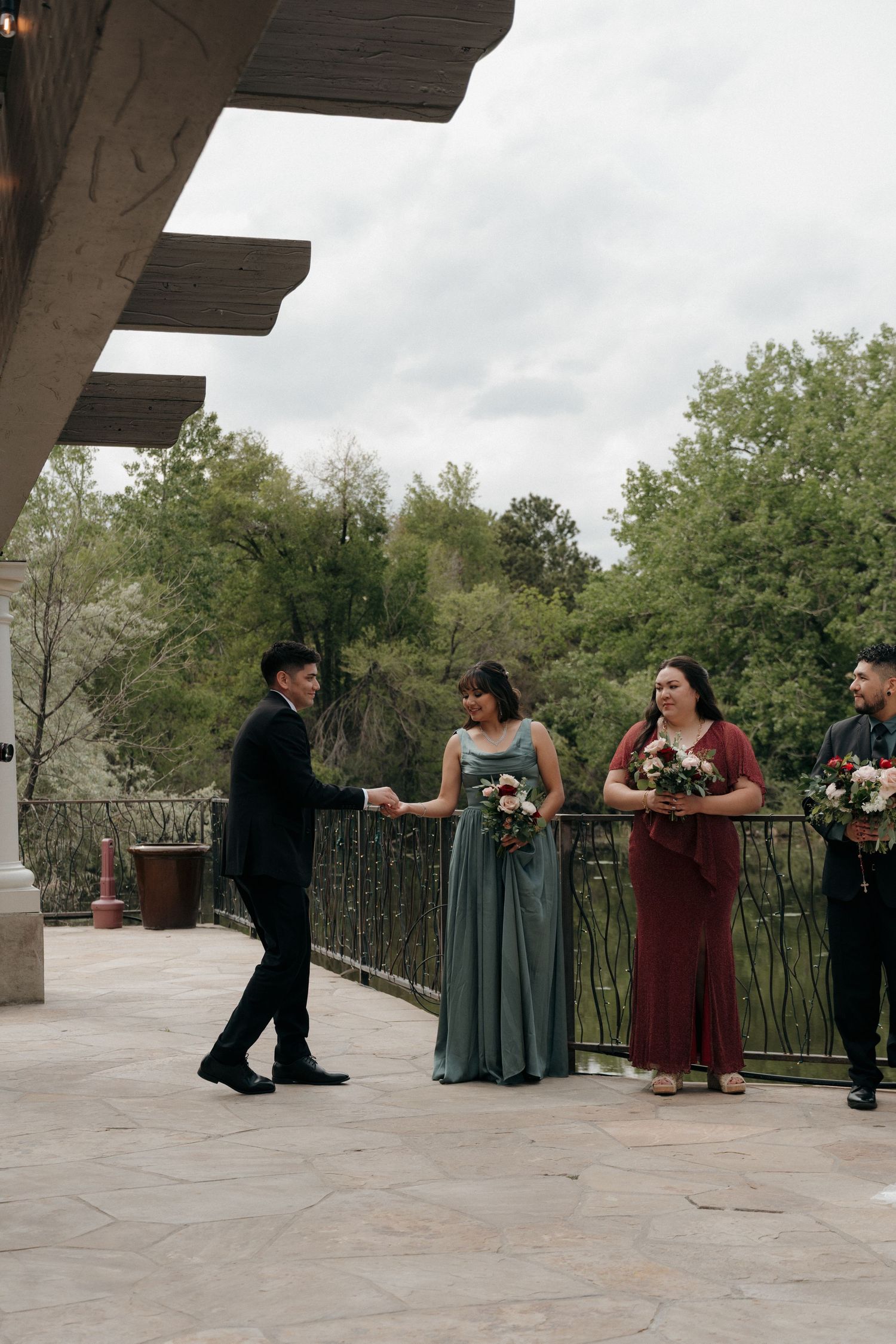 Groom in black suit greets bridesmaids in teal and burgundy dresses on an outdoor venue terrace.