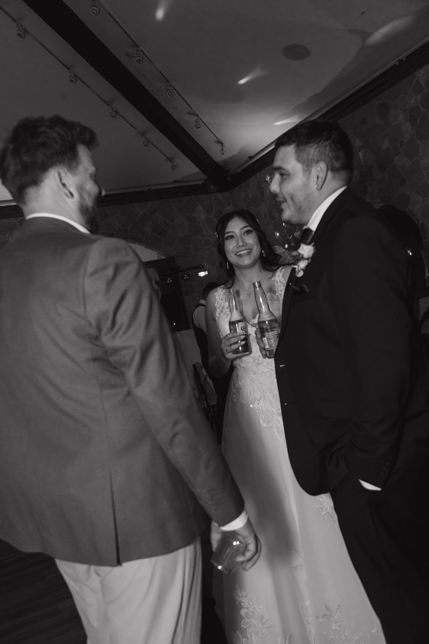 Bride and groom holding champagne glasses chatting with a guest at their wedding reception.