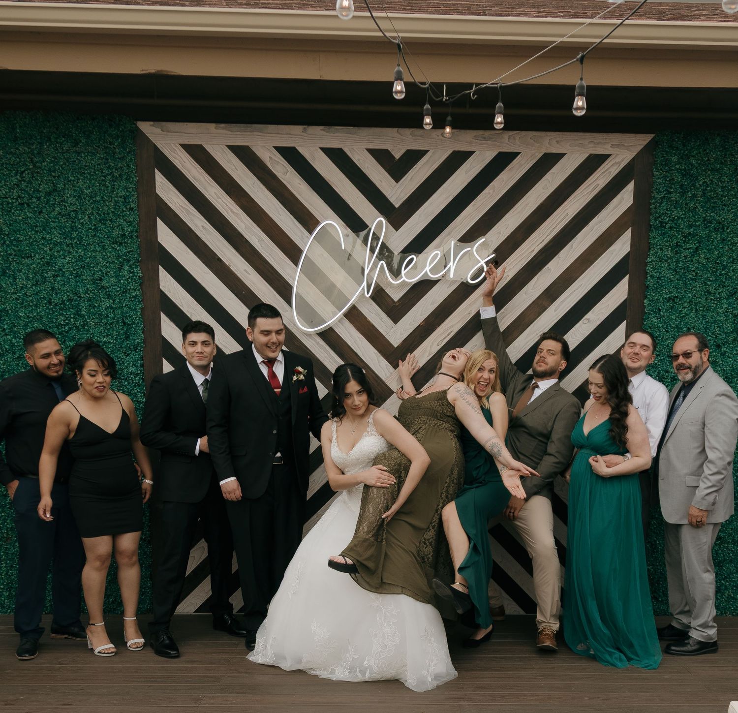 Wedding party posing playfully in front of a striped Cheers sign backdrop at outdoor reception.