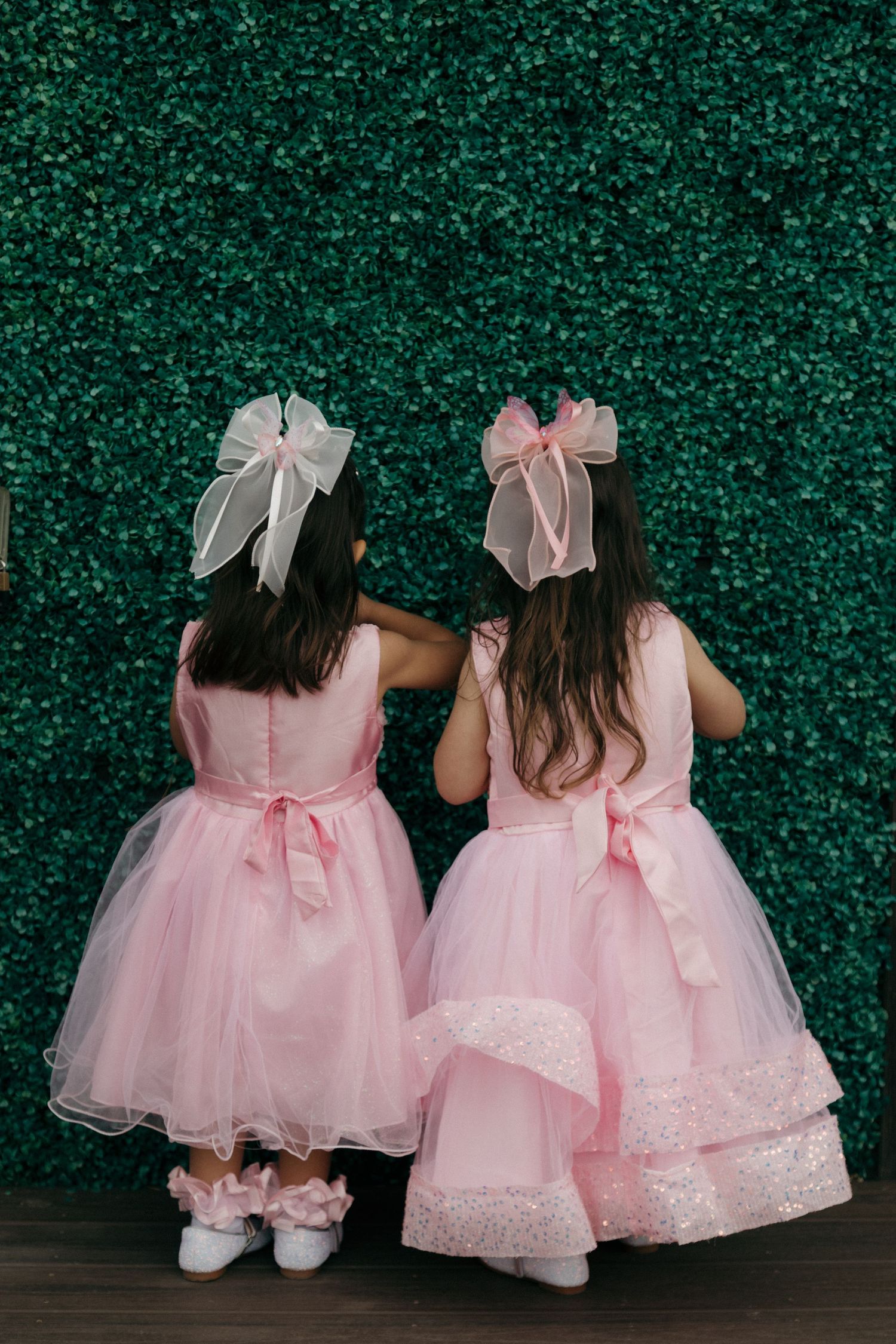 Two little girls in matching pink tutu dresses with white bows pose in front of a green hedge wall.