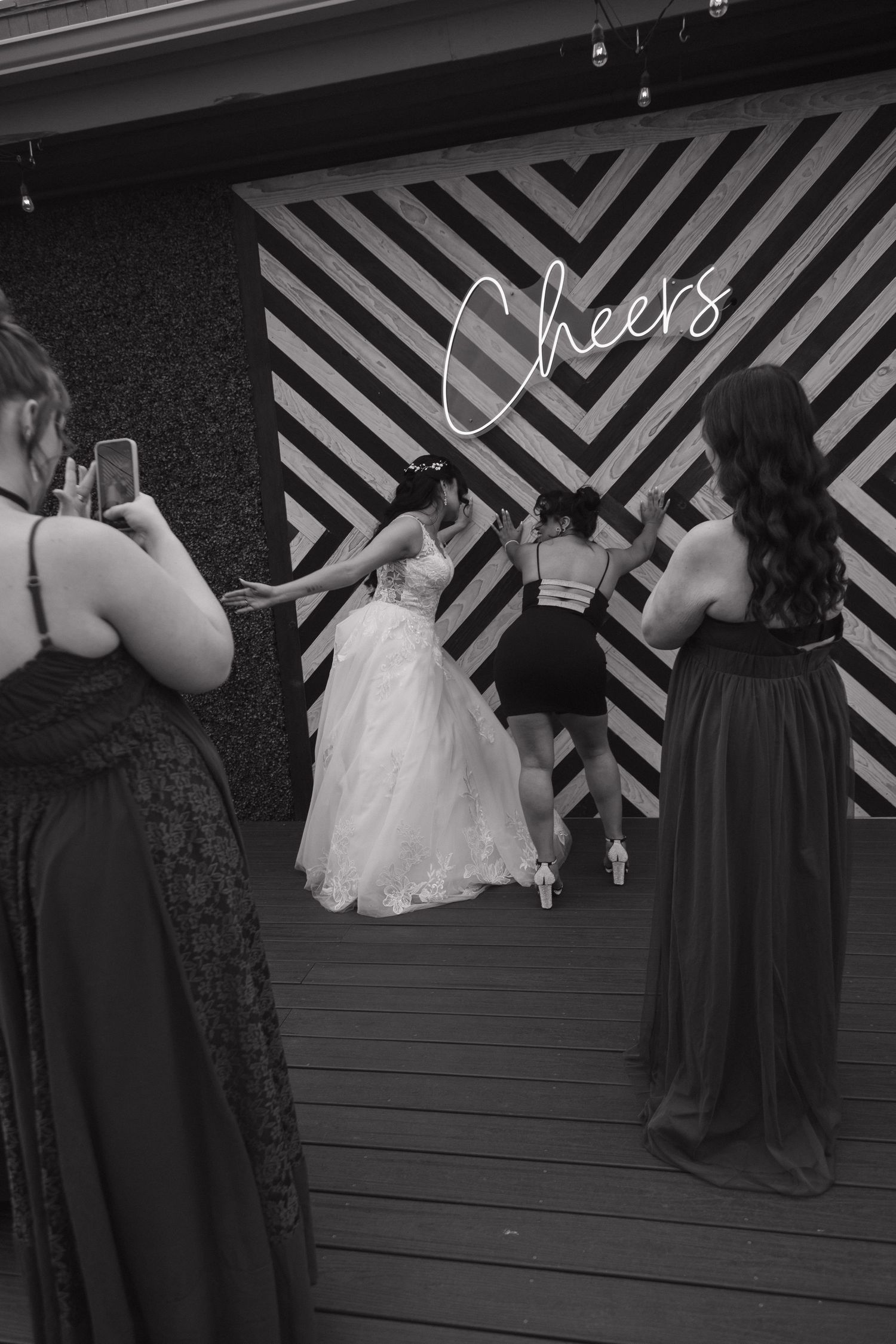 Black and white photo of bride in white gown posing with bridesmaids at chevron Cheers photo backdrop.
