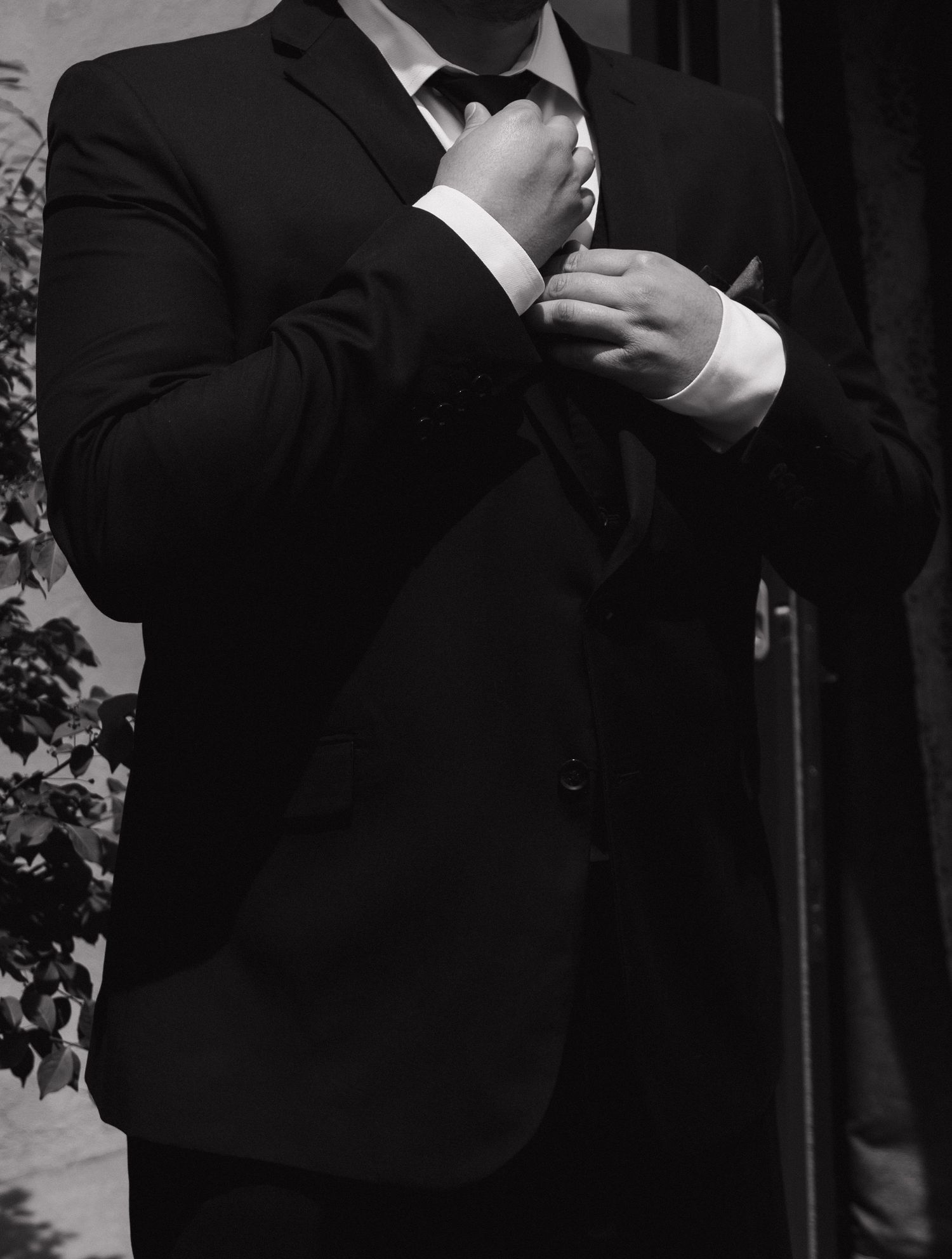 Black and white photo of groom adjusting tie in formal black suit.