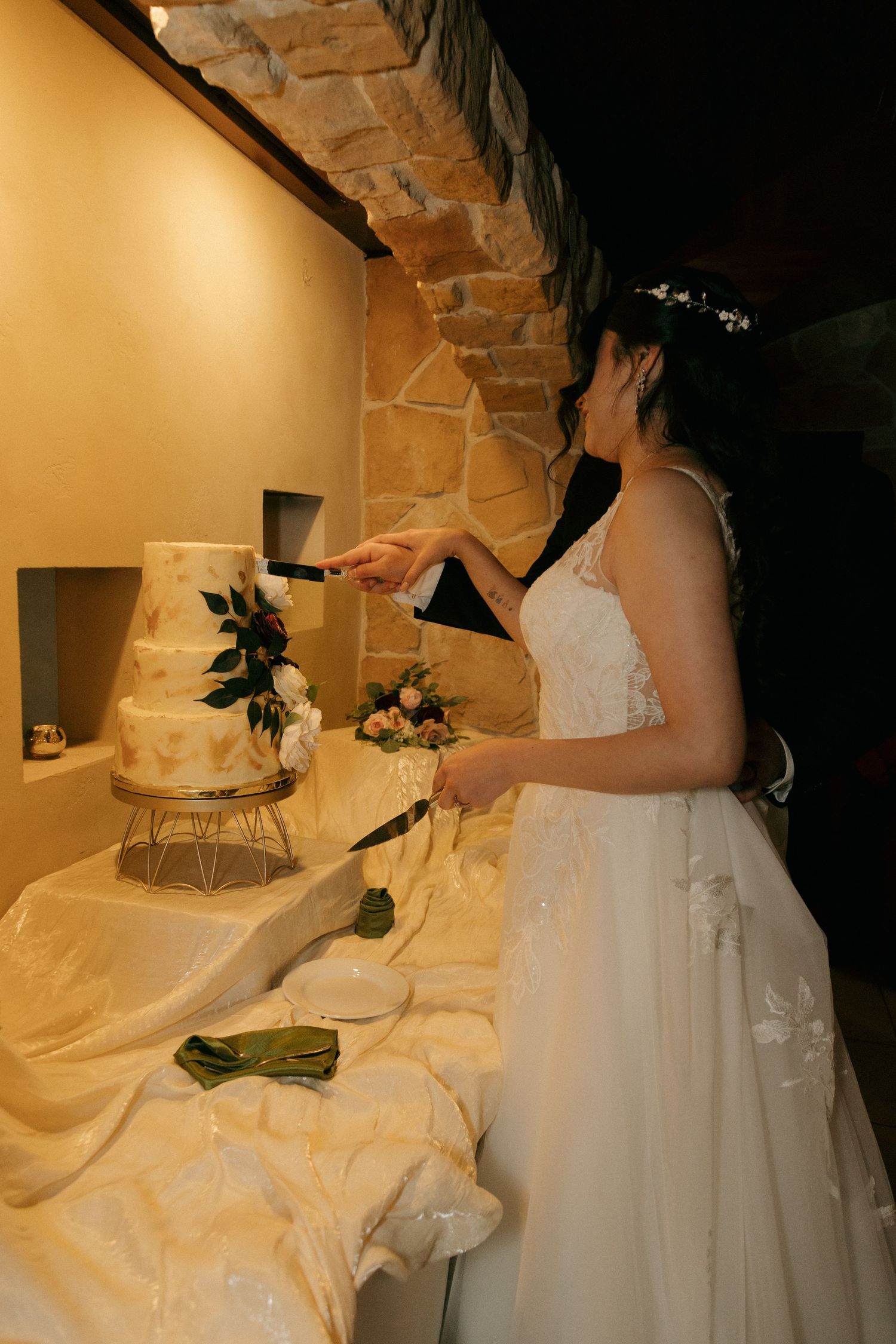 Bride cutting a three-tiered white wedding cake decorated with flowers at the reception.