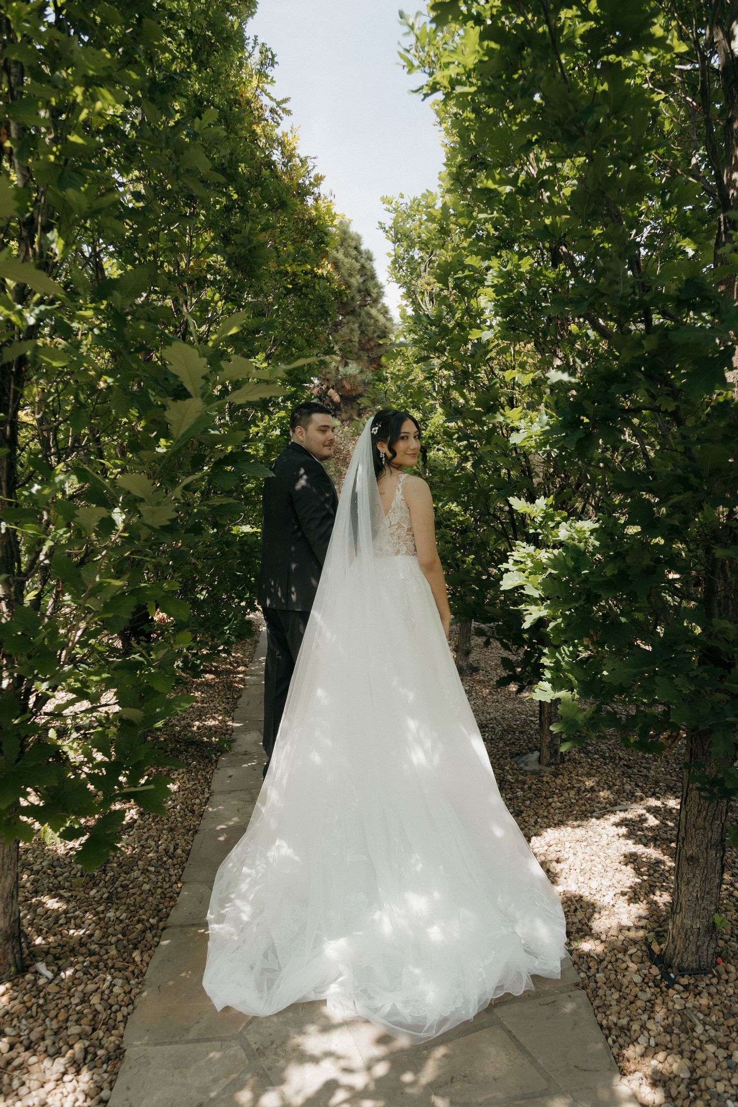 Bride and groom posing on a garden pathway lined with lush green trees.