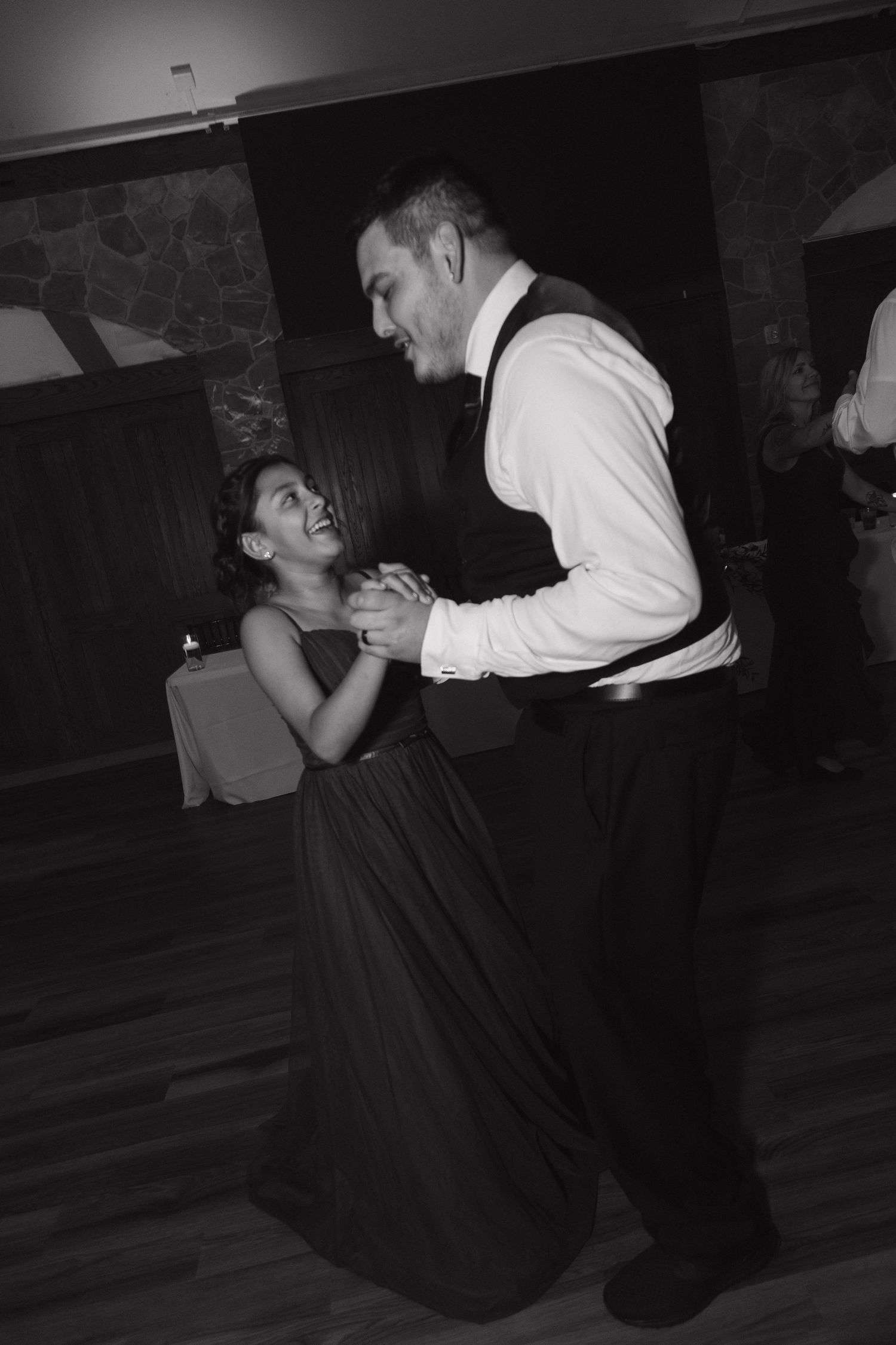 Groom dances and smiles with a young girl in a dark gown at a wedding reception in black and white.