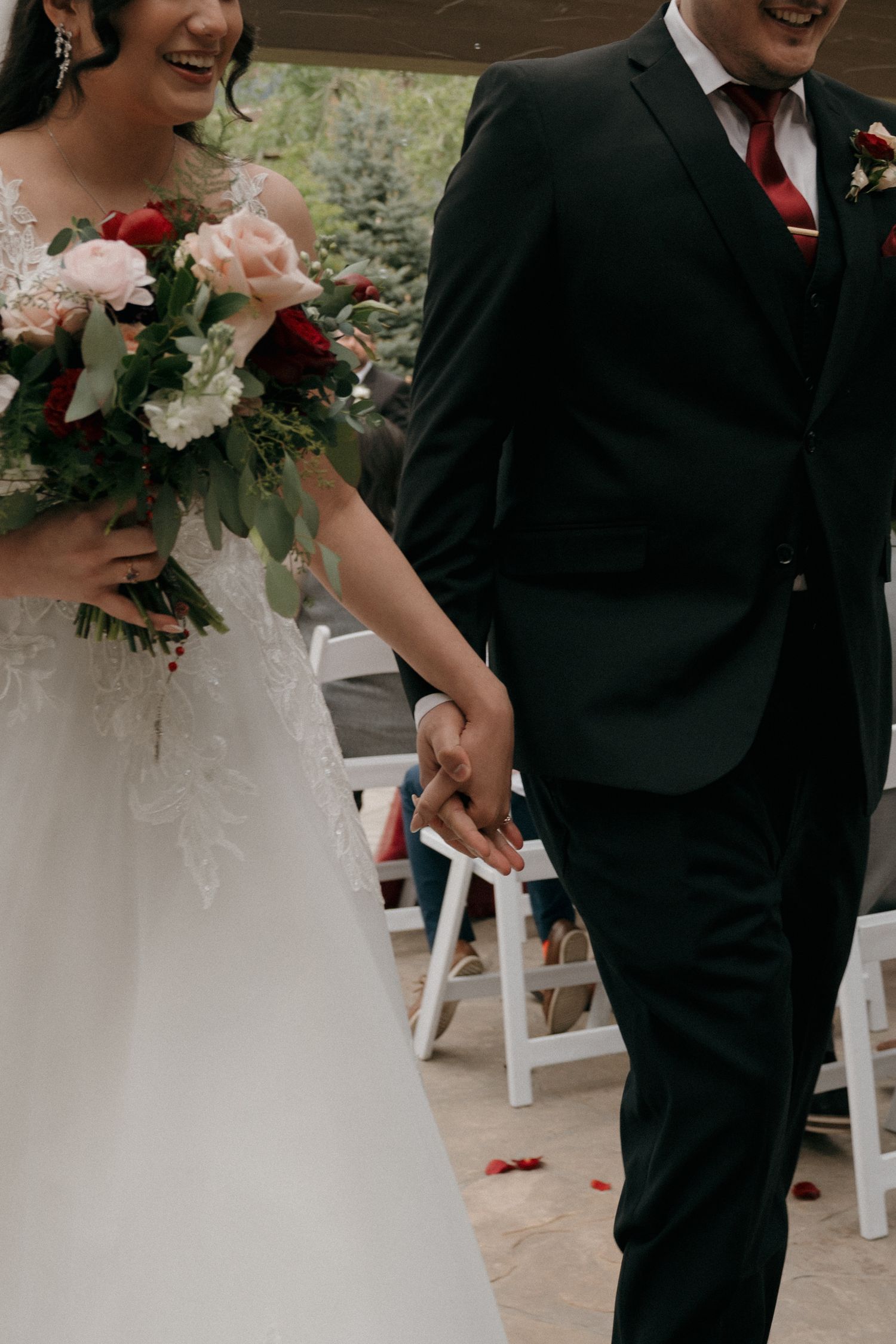 Newlyweds walk hand in hand down aisle, bride holding red and white floral bouquet.
