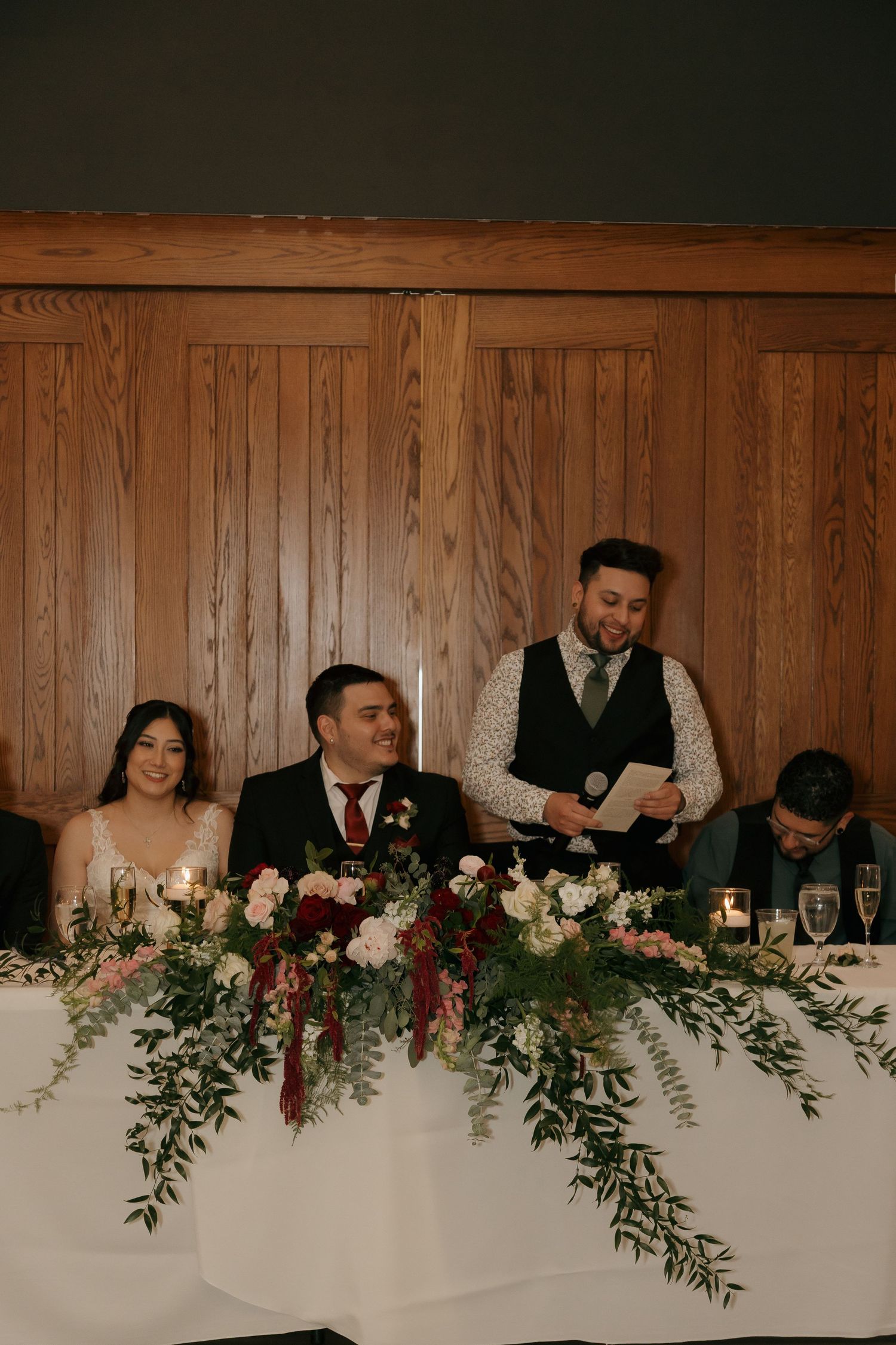 Best man reads toast as bride and groom smile at floral-decorated head table during wedding reception.