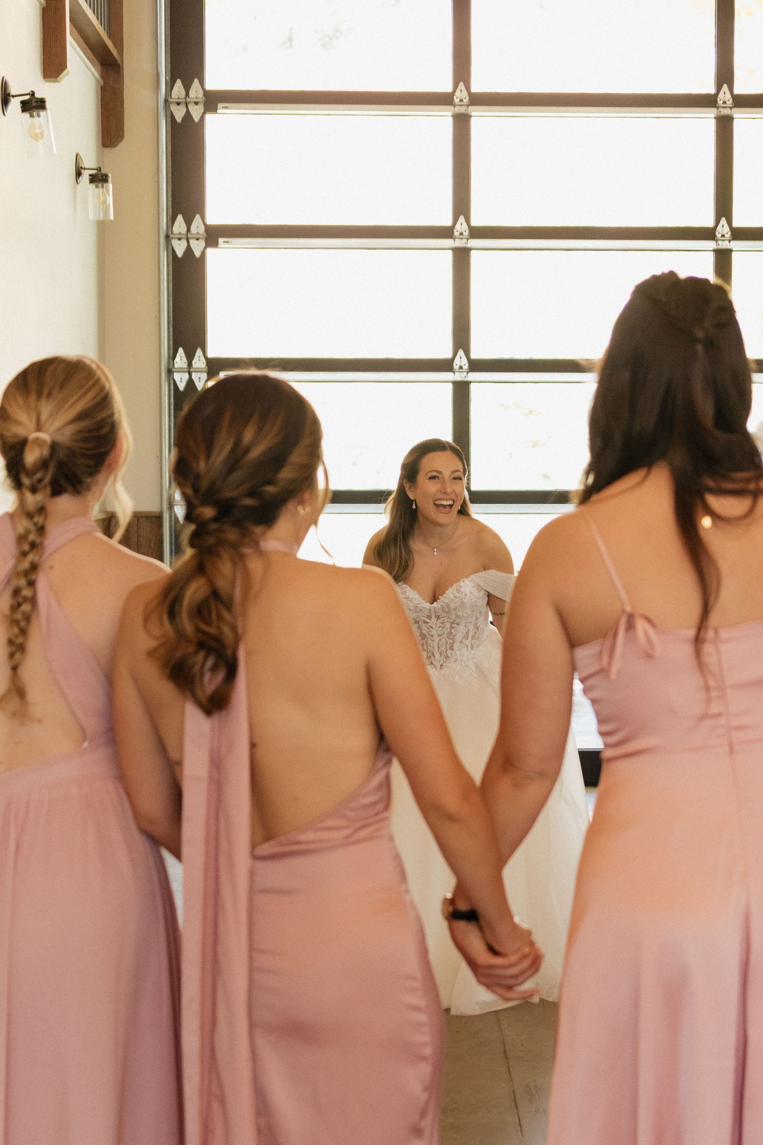 A row of bridesmaids in pink dresses stand together looking out windows in a wedding preparation room.