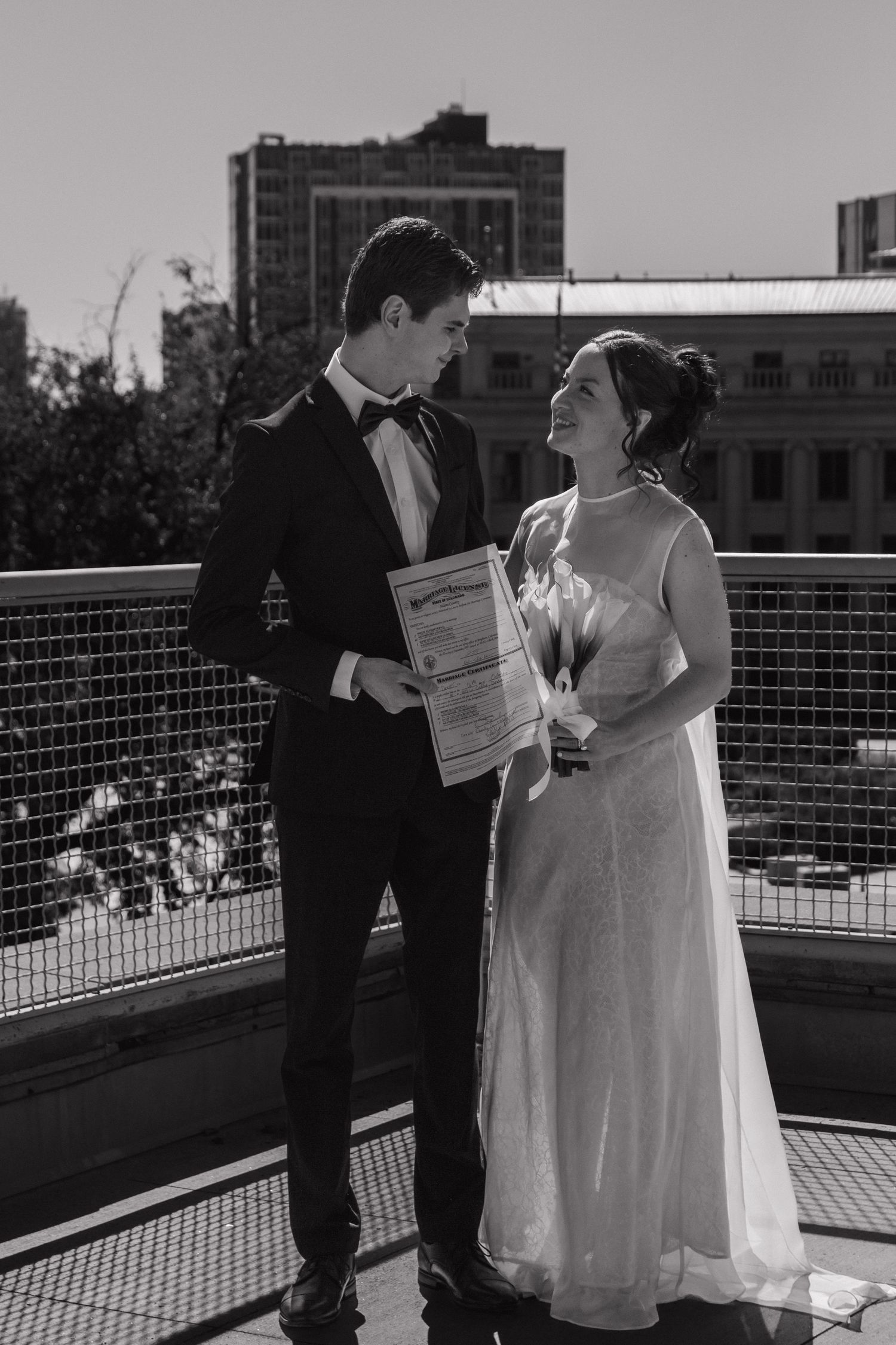 Black and white photo of a couple exchanging vows on a rooftop with city buildings in the background.