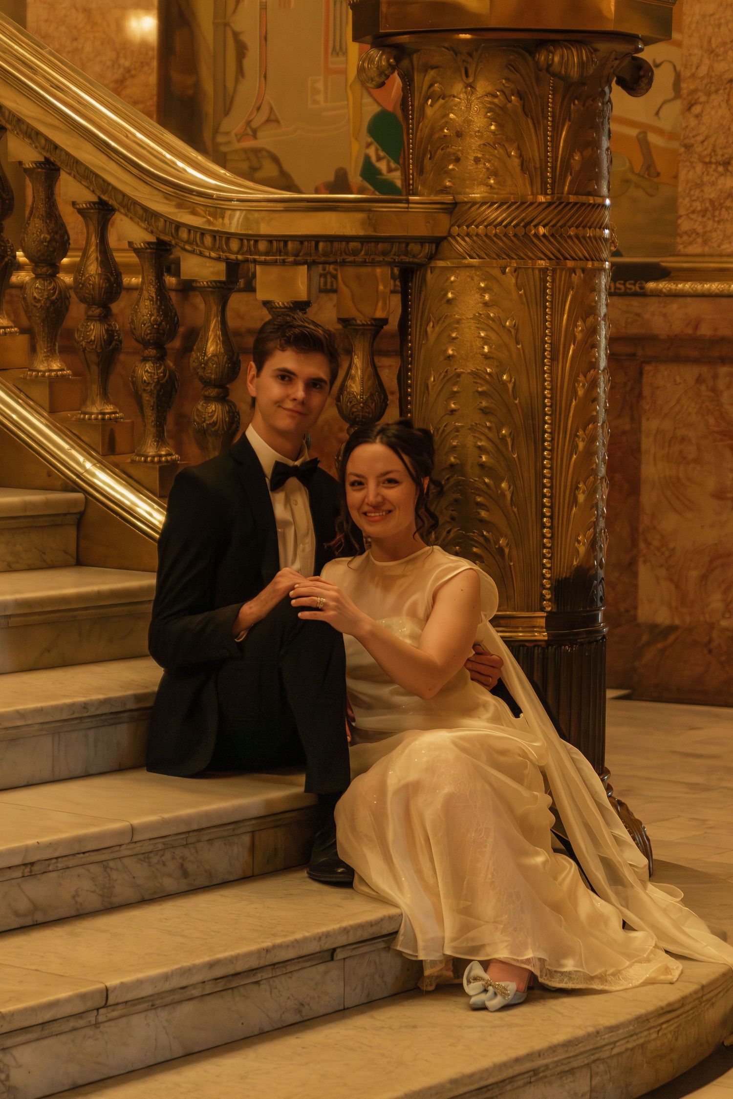 A couple in formal attire sits on an elegant marble staircase with ornate gold railings and decorative columns.
