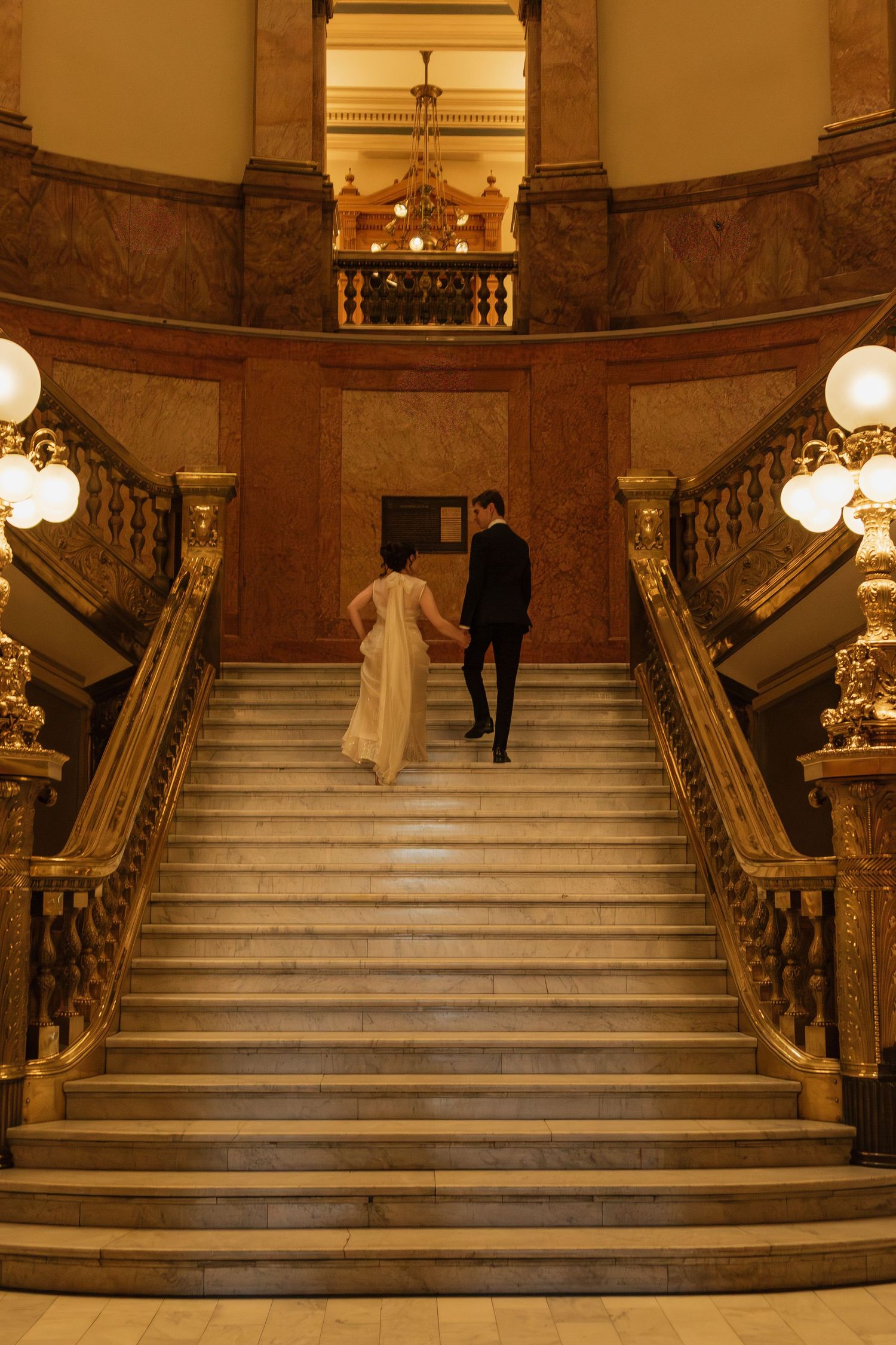 Grand marble staircase with ornate gold railings and glowing globe lights in an elegant historic building interior.