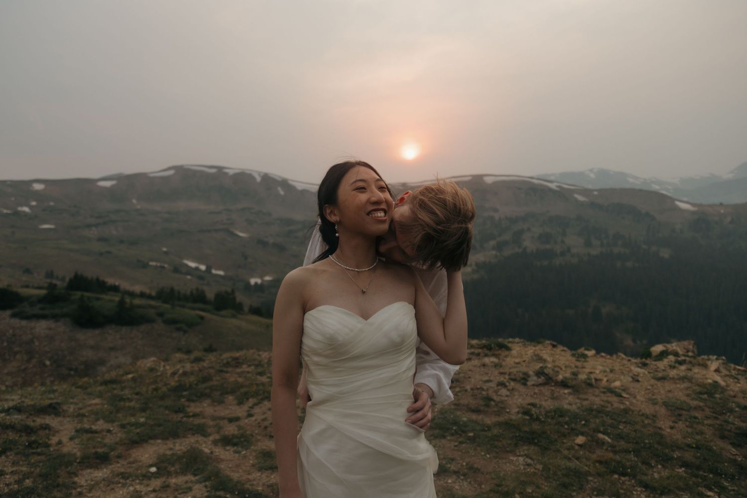 Groom kisses bride on the cheek at sunset with smoky mountain panorama behind them.