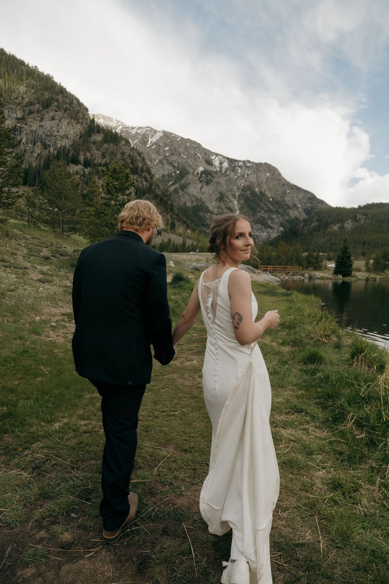 Bride in white lace-back gown holds groom's hand walking near mountain lake.