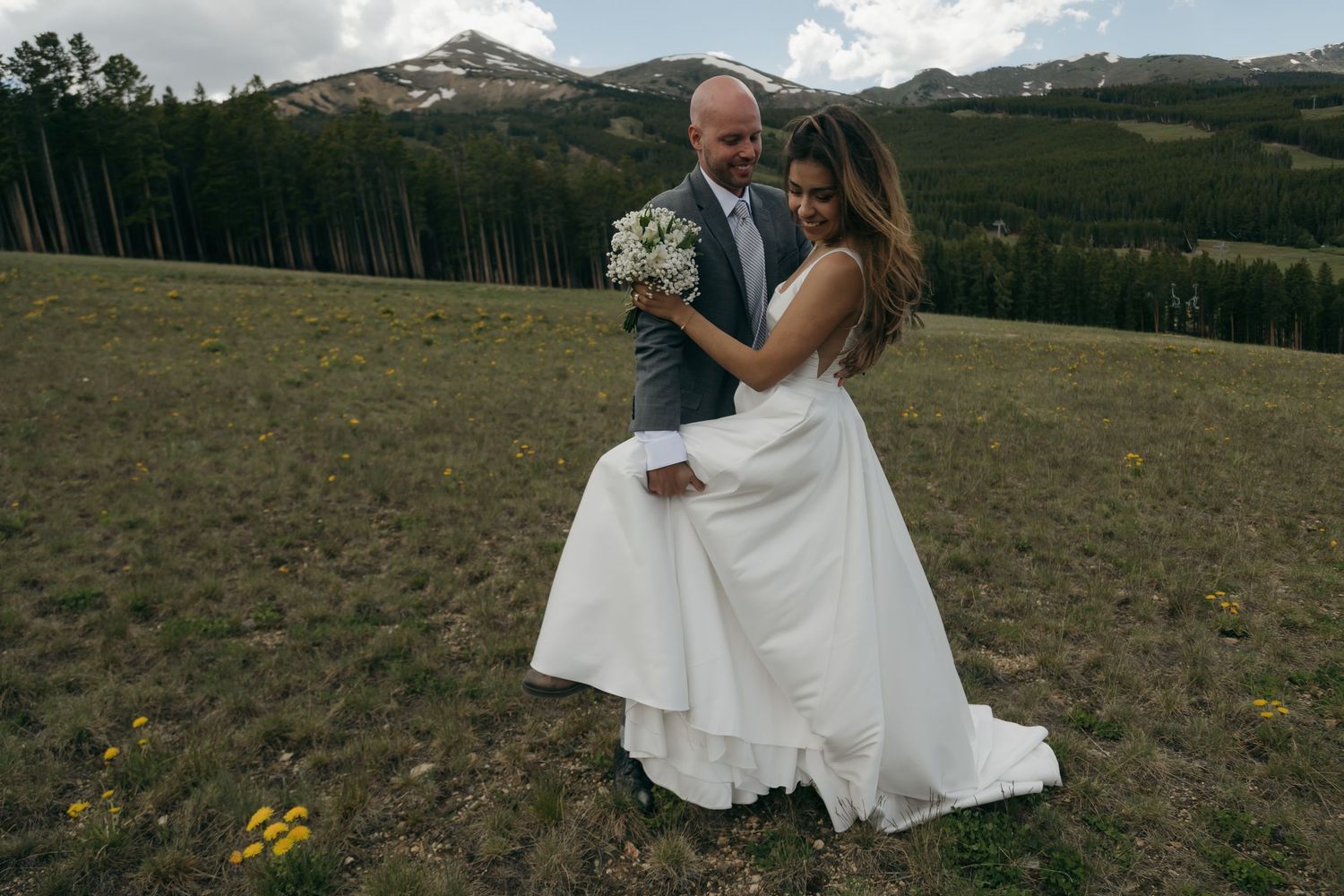 Bride and groom embrace in a mountain meadow, bride holding white bouquet in elegant white gown with forest backdrop.