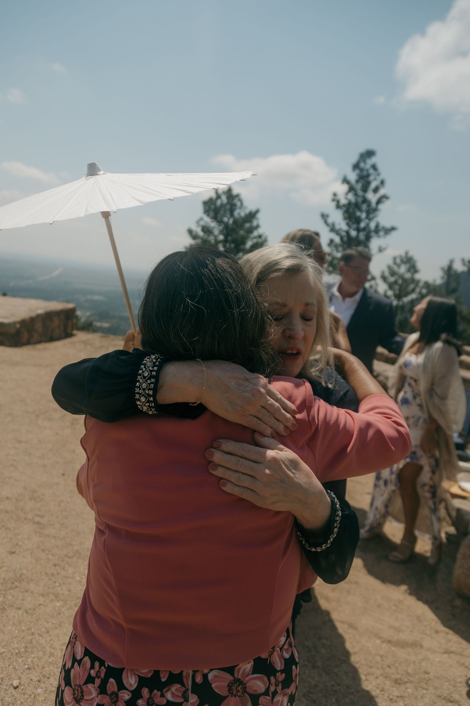 Two women sharing an emotional hug outdoors at a mountain wedding ceremony under sunny skies.