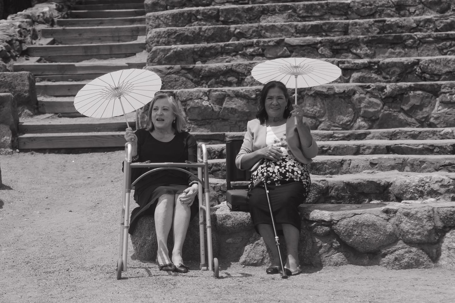 Two women sit in chairs holding white parasols near stone steps at an outdoor wedding ceremony.
