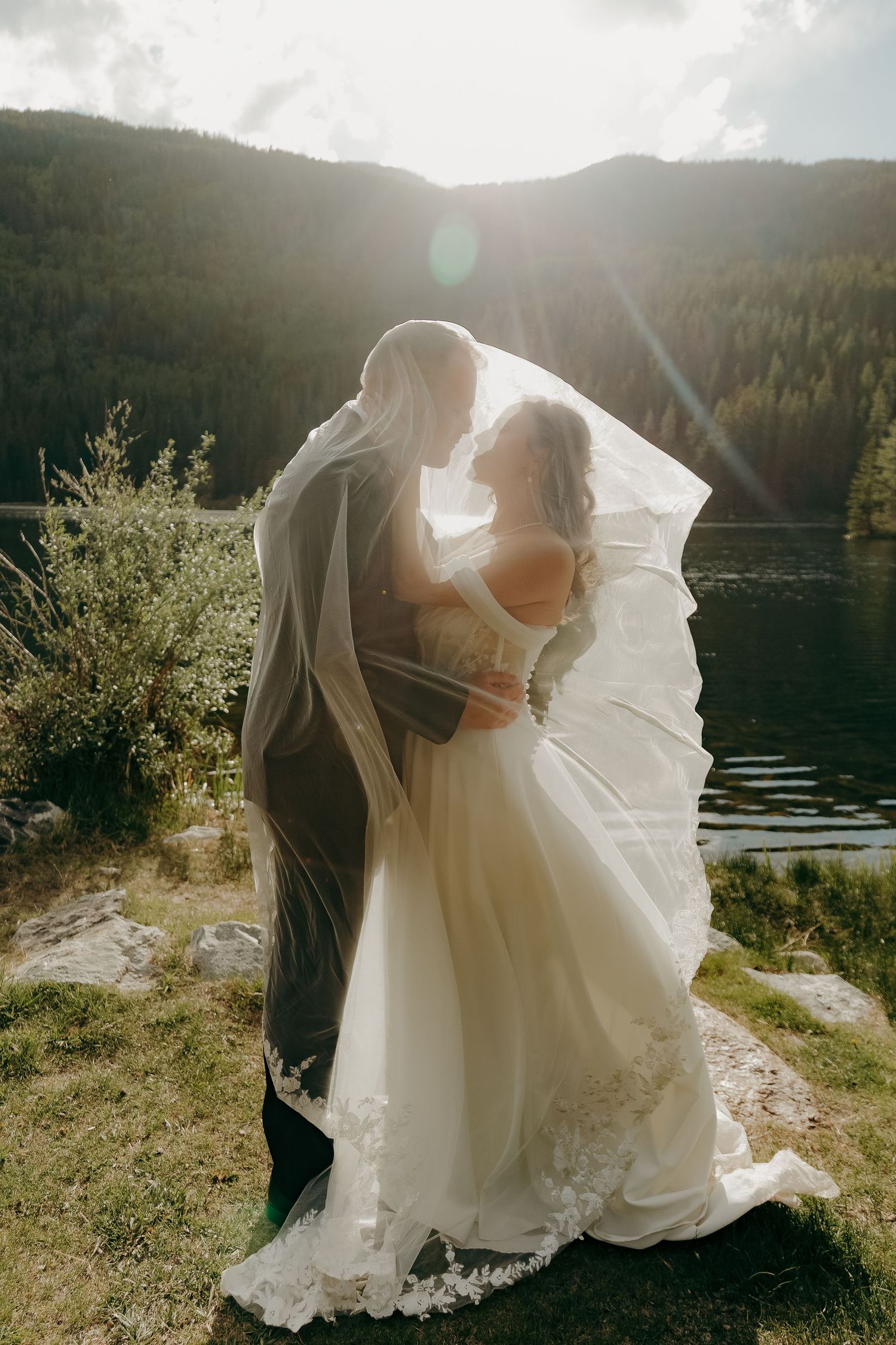 Bride and groom sharing a romantic moment under a flowing veil by a mountain lake in golden sunlight.