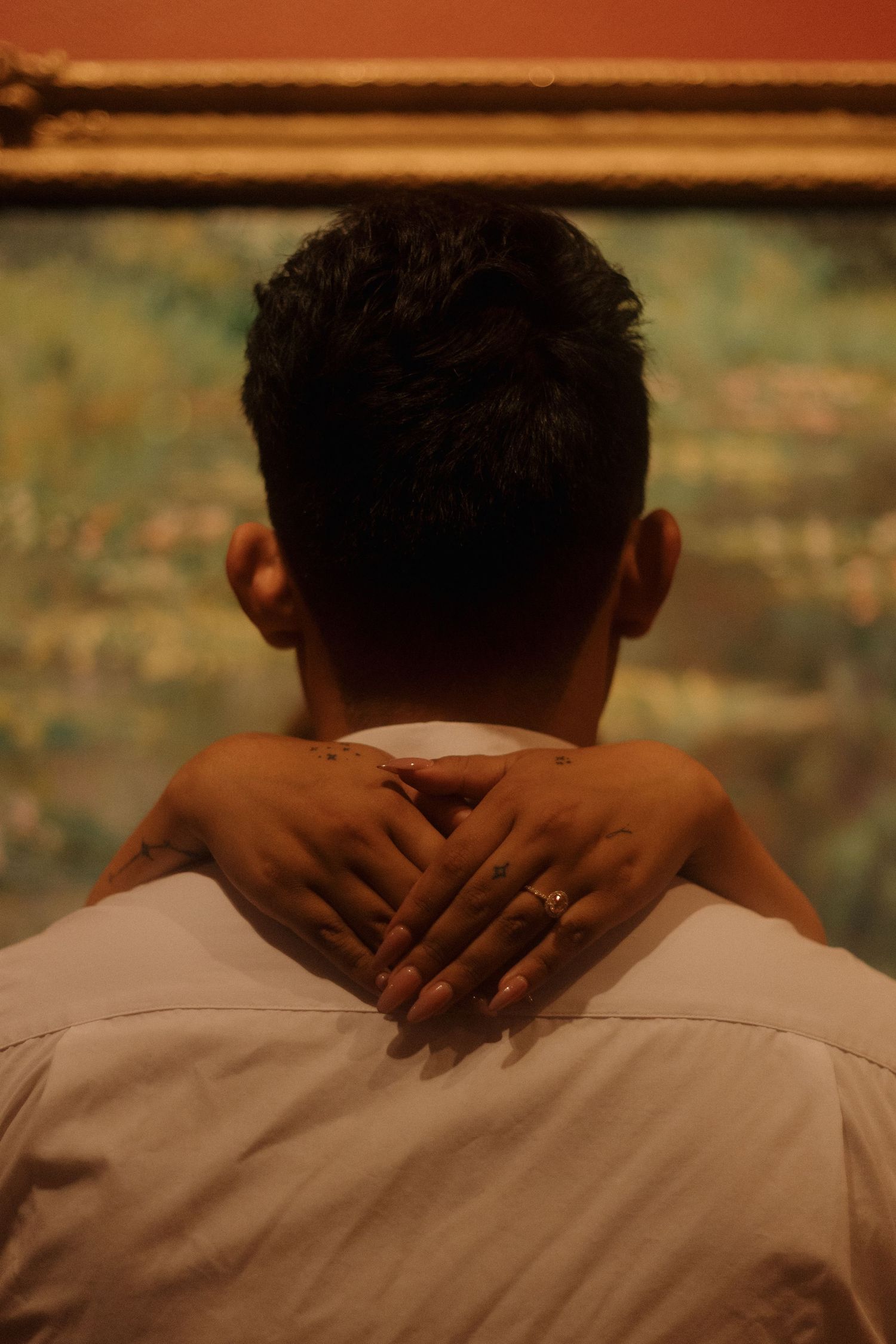 Back view of a person being embraced, hands with a ring resting on shoulders, standing before a impressionist painting.