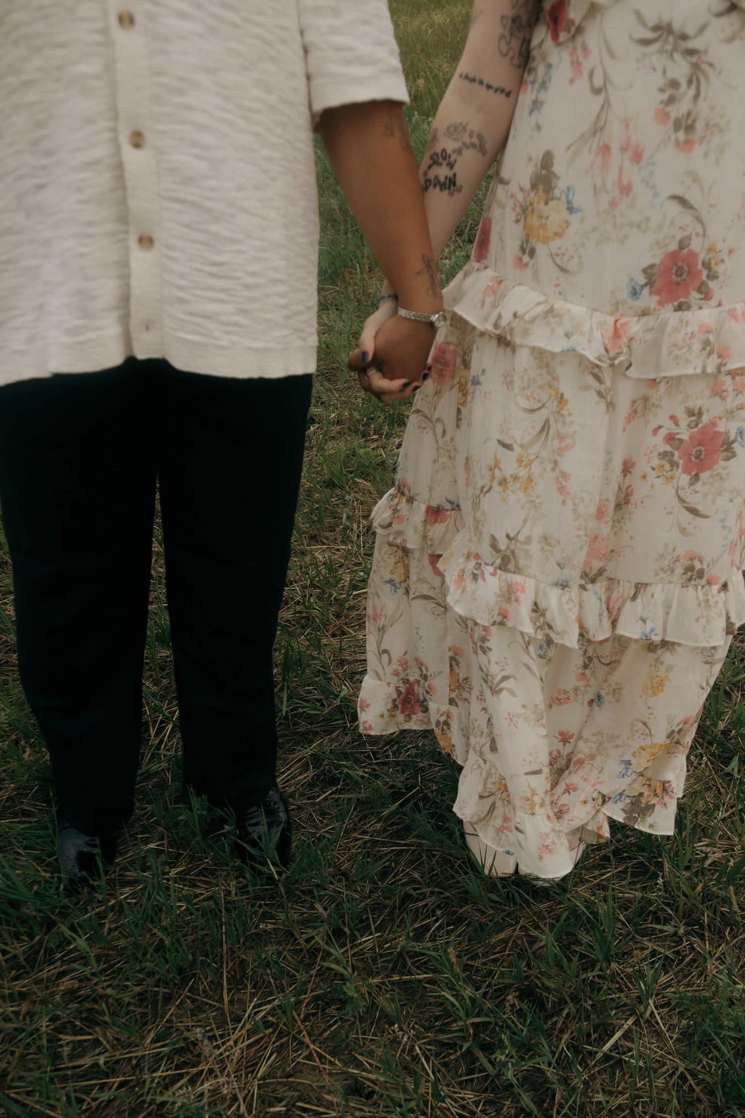 Couple holding hands in a field, woman wearing a floral ruffle dress with tattoos, man in black pants.