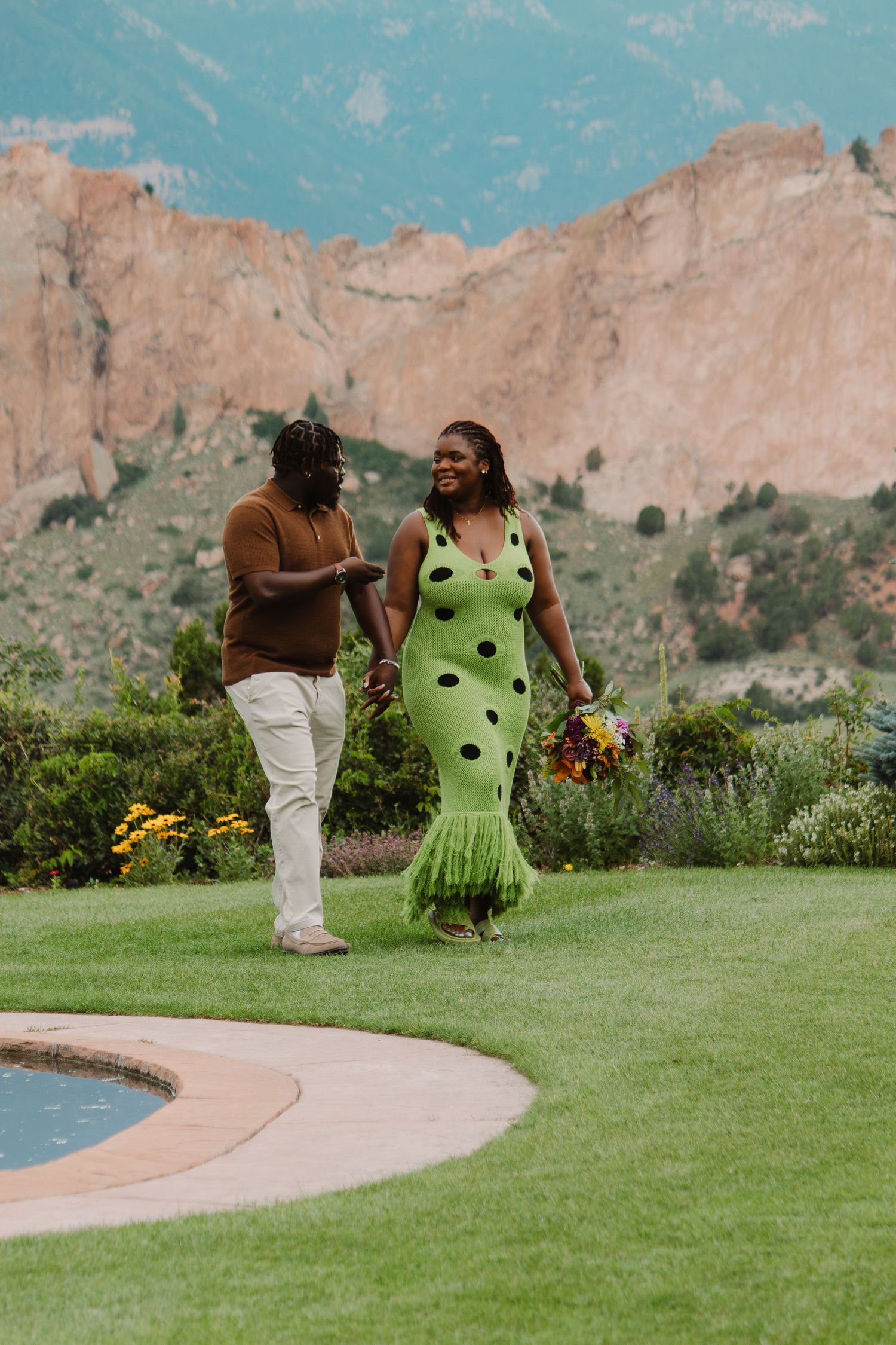 Couple in contrasting outfits sharing a romantic moment on green lawn with mountain backdrop.