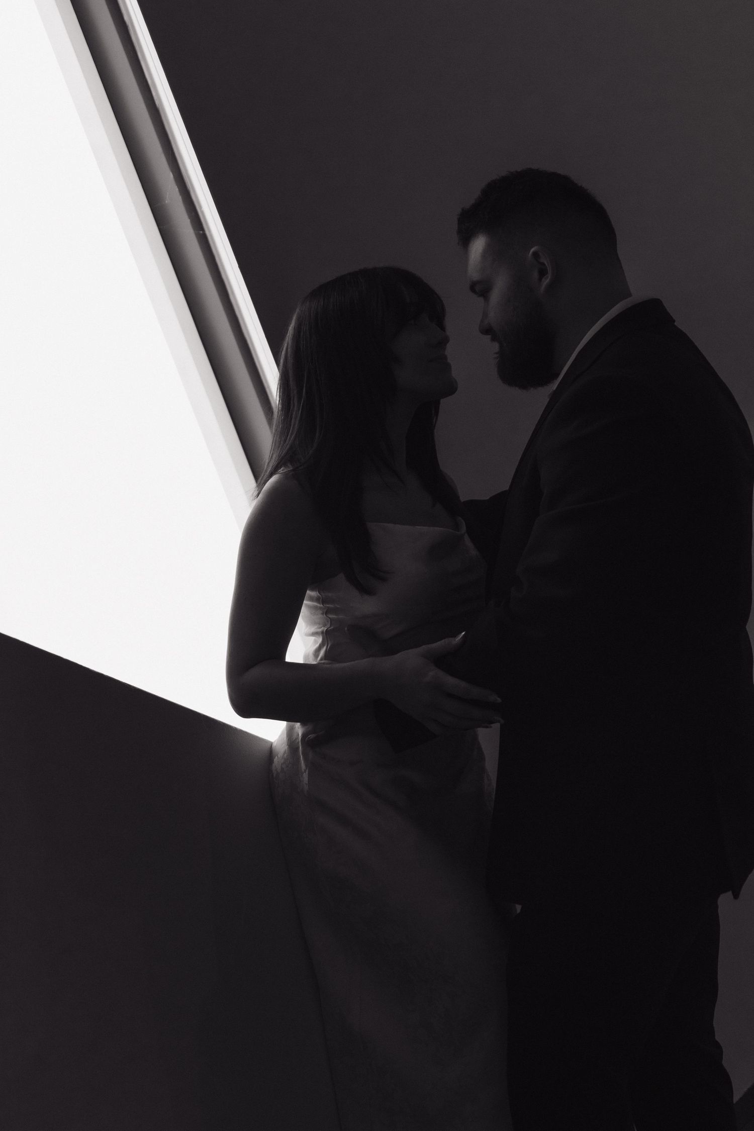 A silhouetted couple in formal attire share an intimate moment by a window in dramatic black and white lighting.