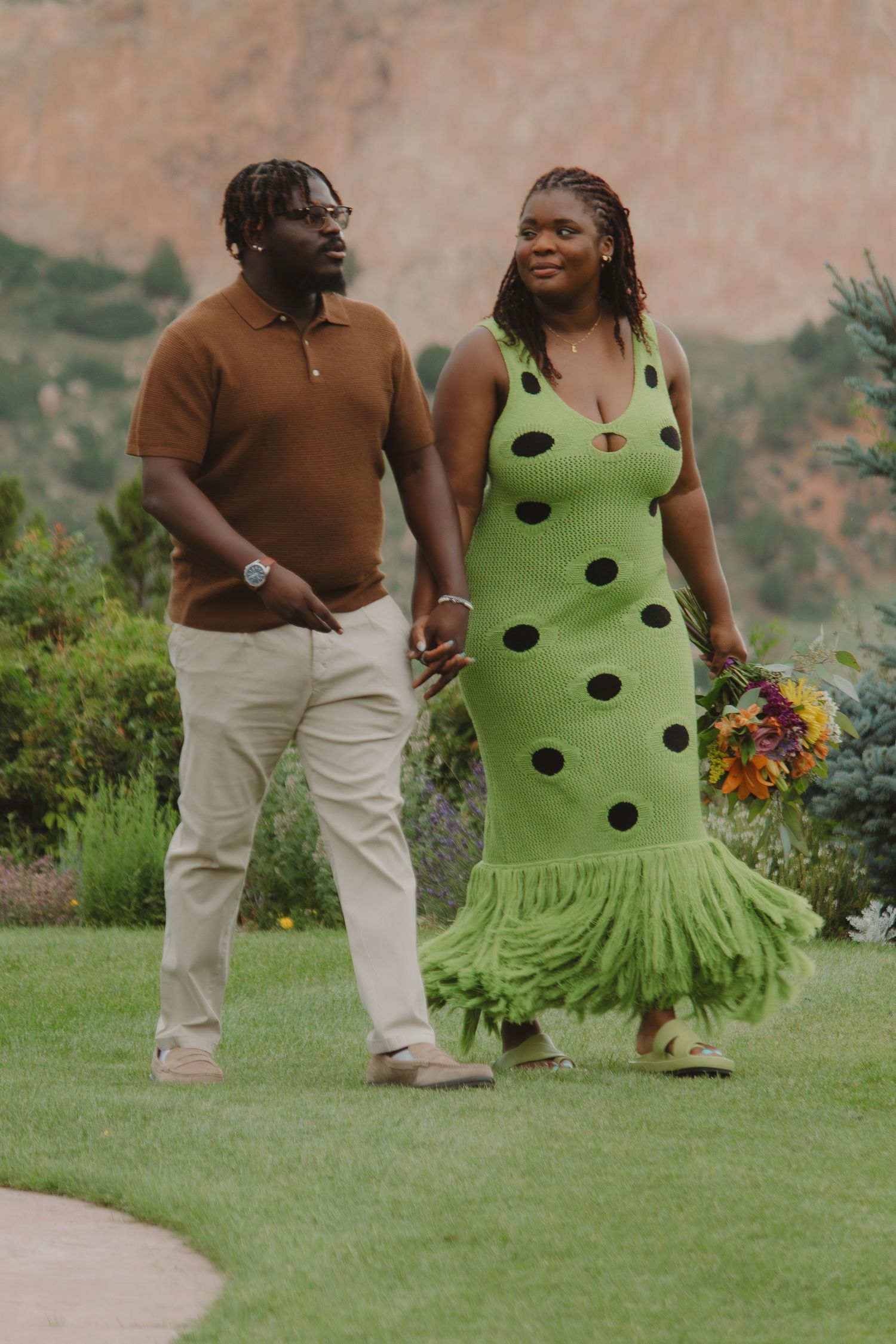 Couple strolls through a green garden path, one wearing a polka dot dress with ruffled hem.