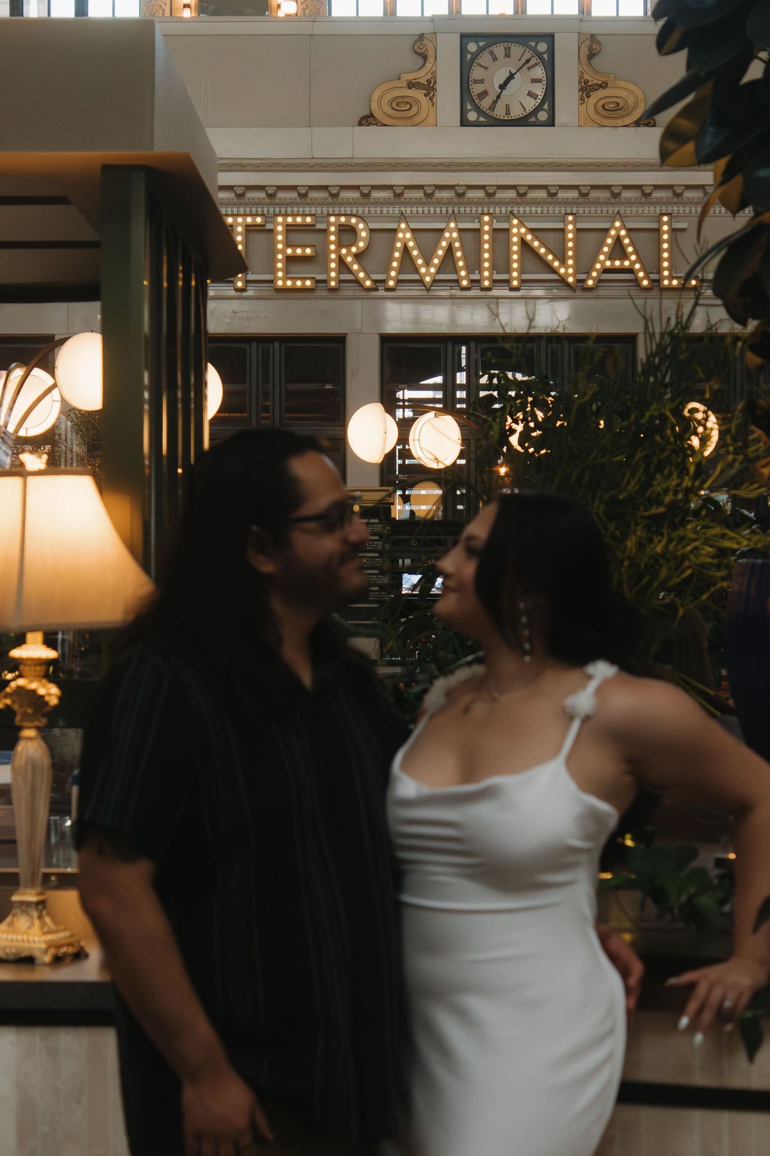 A romantic moment under illuminated TERMINAL signage with vintage-style lighting and indoor plants.