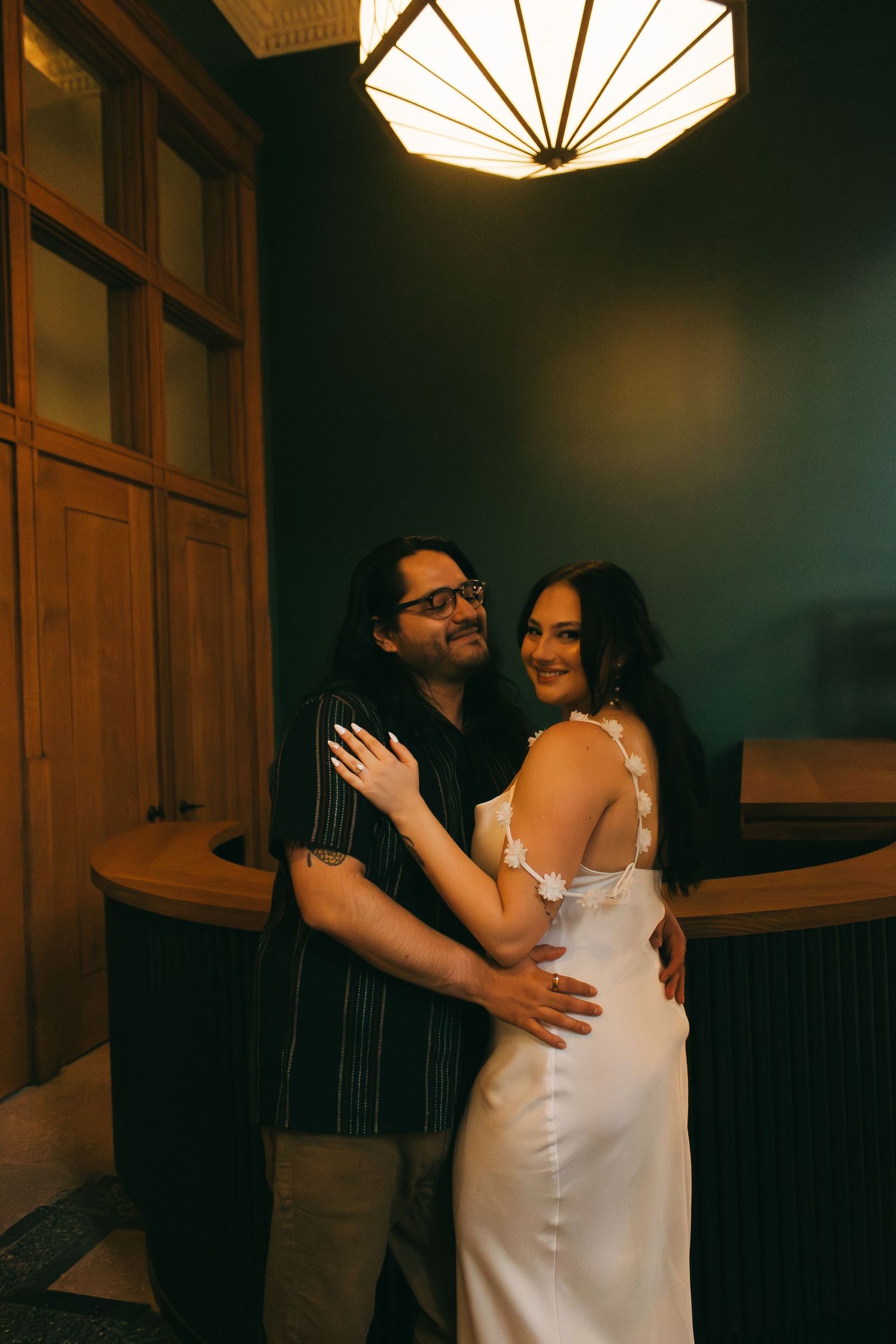 Couple embraces in front of decorative ceiling light fixture during indoor photoshoot.
