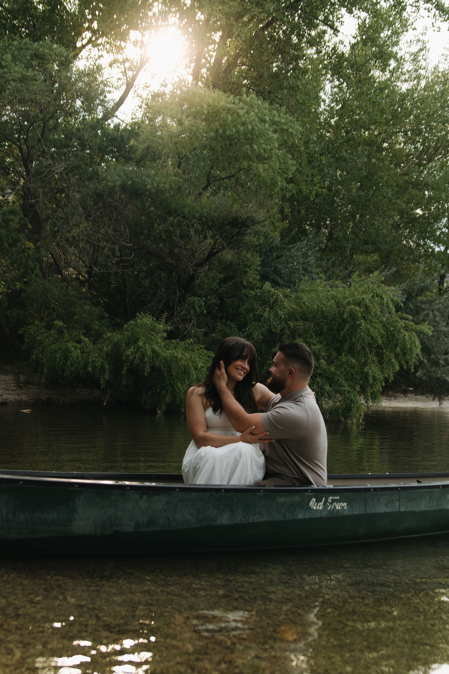 Couple cuddling together while sitting in a dark green canoe surrounded by overhanging trees.