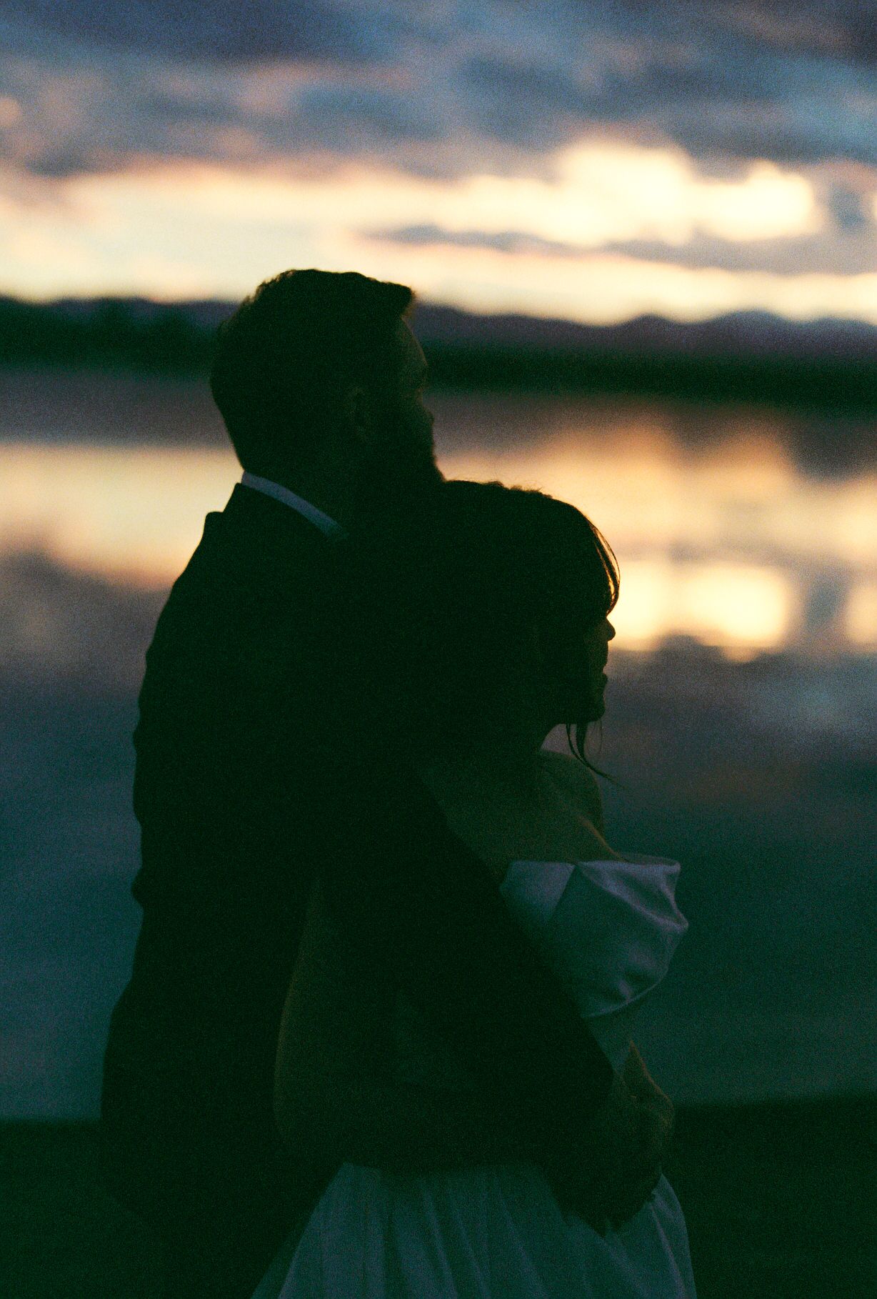 Silhouetted newlyweds embrace during golden hour against a dramatic sky.