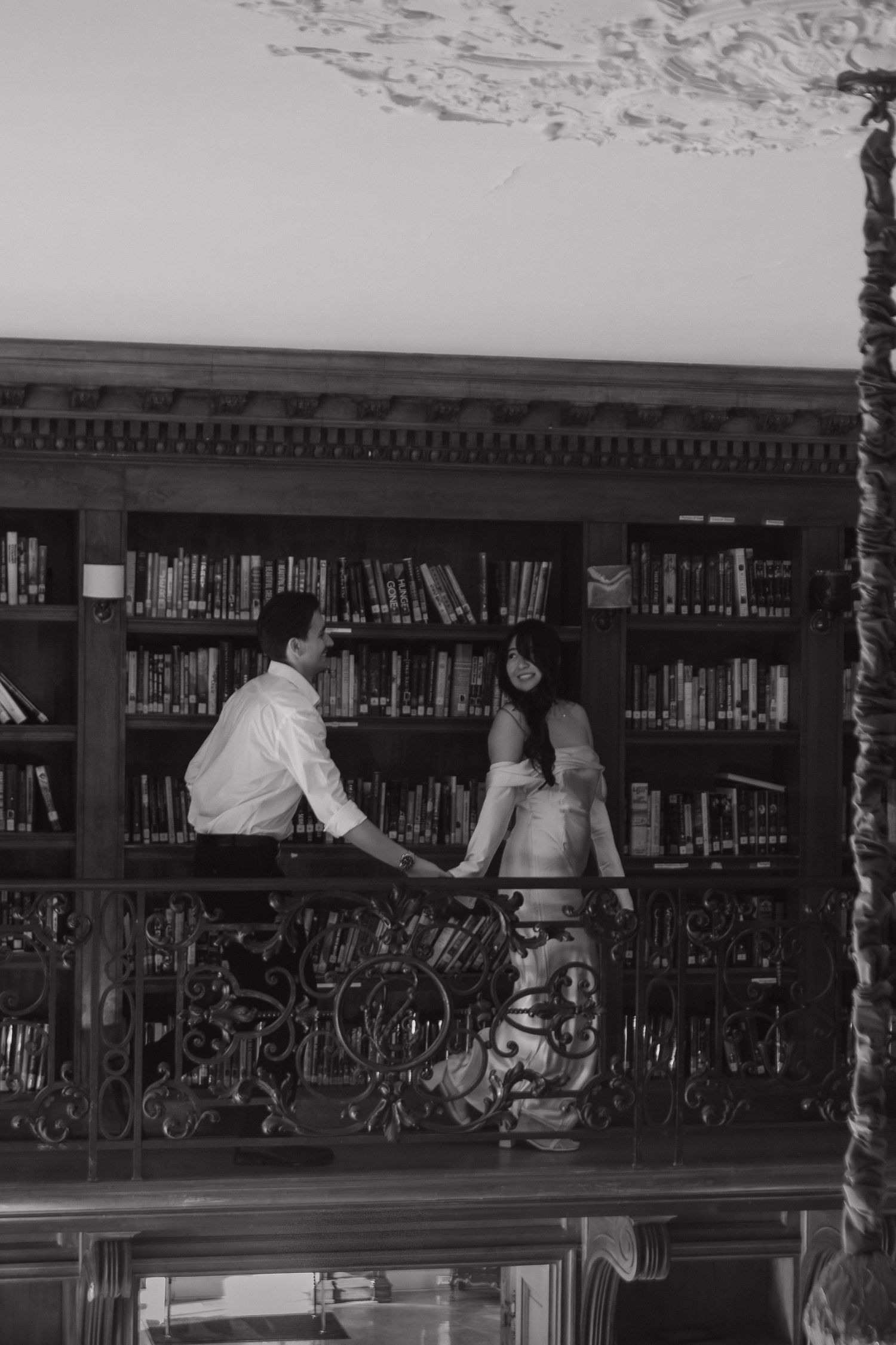 A couple embraces romantically in a vintage library with ornate iron railings and wooden bookshelves.