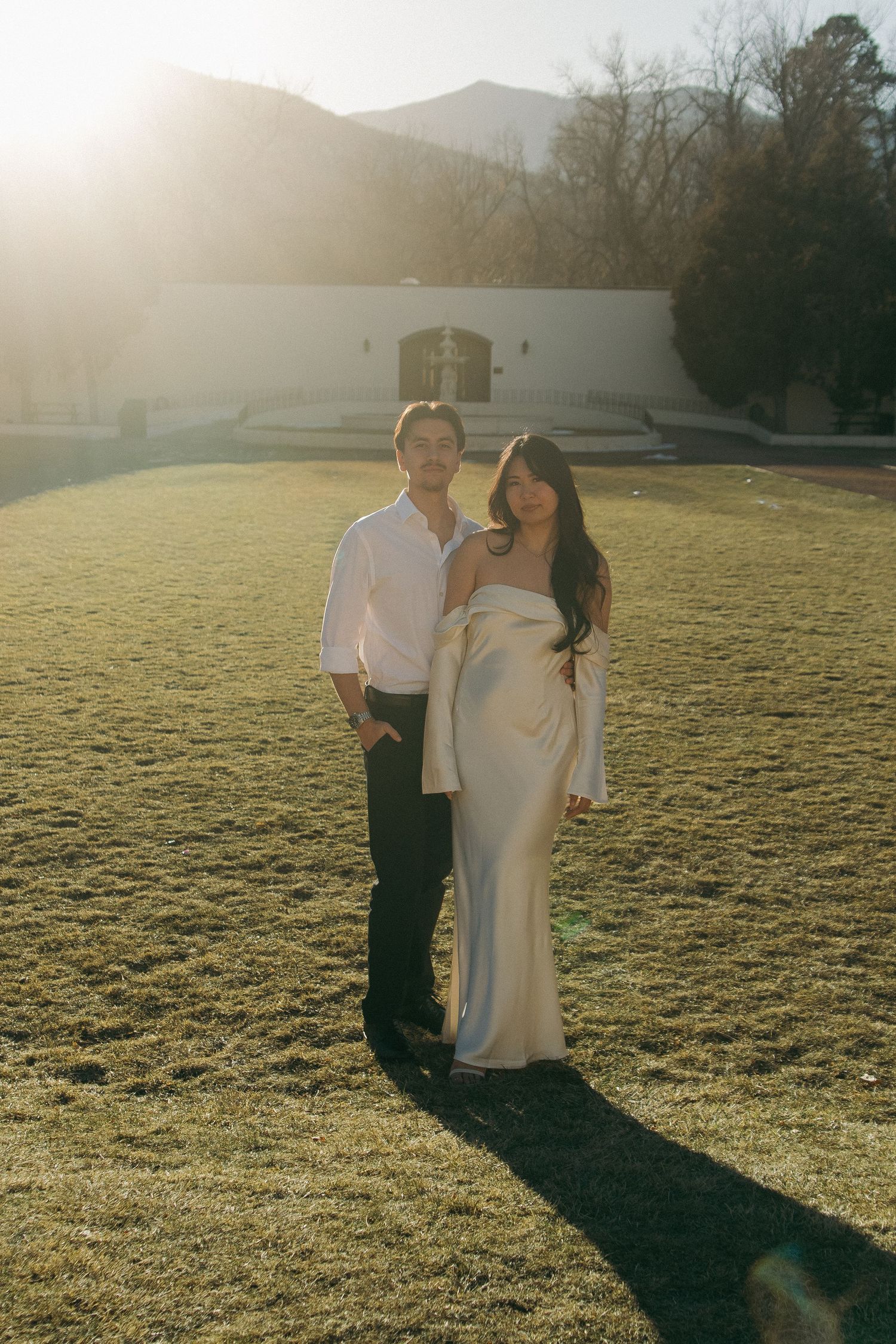 A couple poses together on a sunlit lawn with mountains and a white building in the background.
