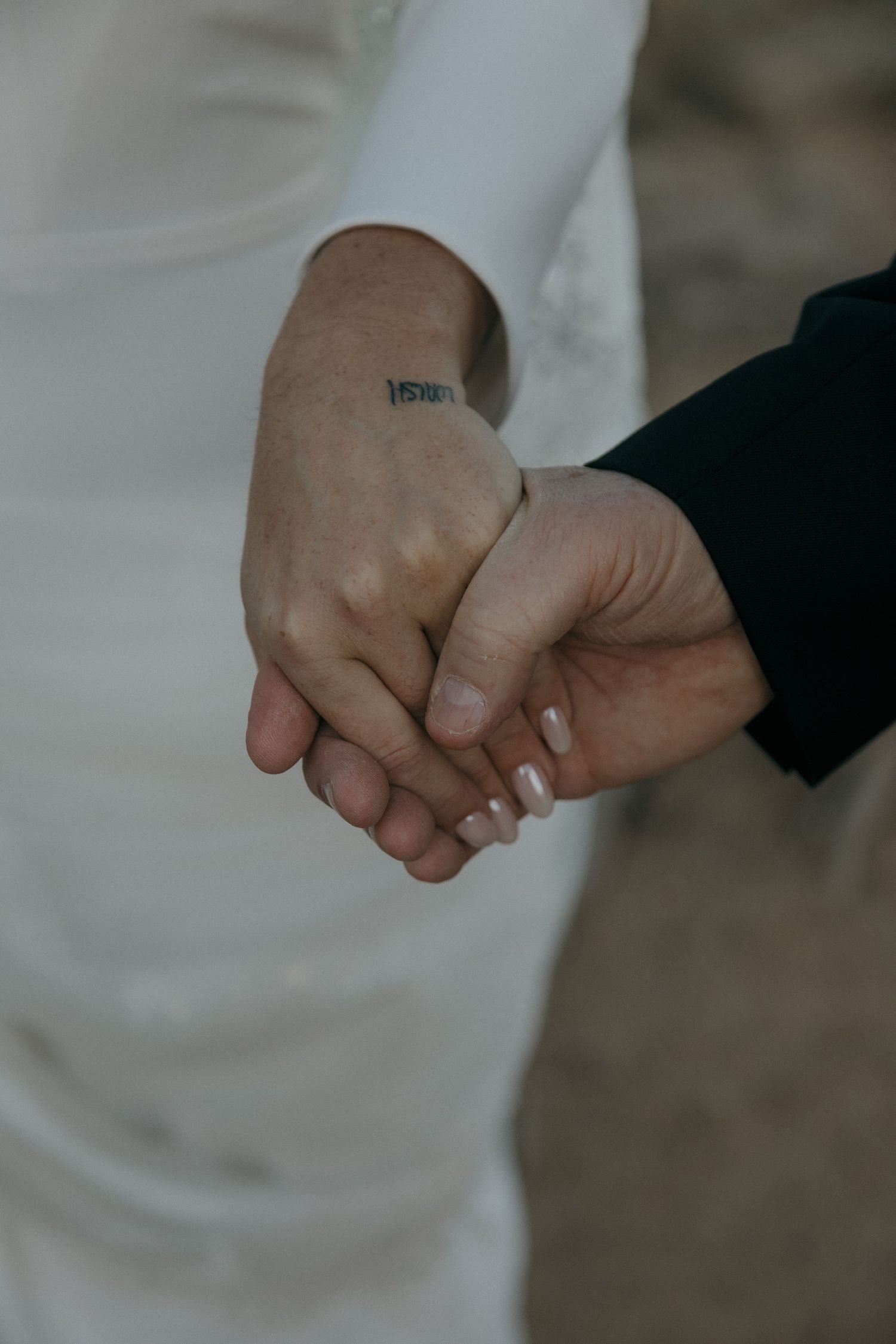 Close-up of bride and groom holding hands, bride wearing white dress with small wrist tattoo.