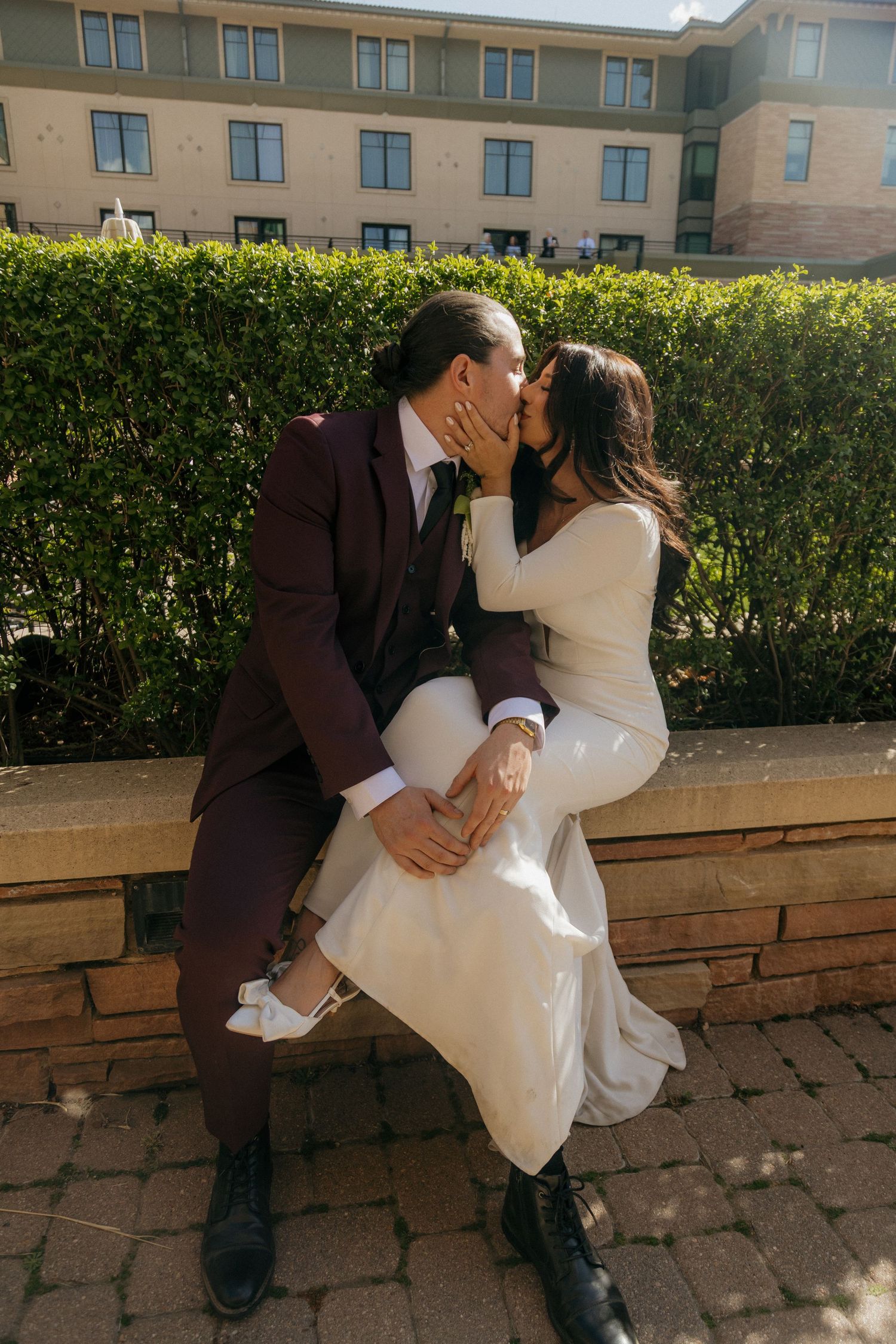 Bride and groom sharing a kiss while seated on a brick ledge in front of a green hedge outdoors.