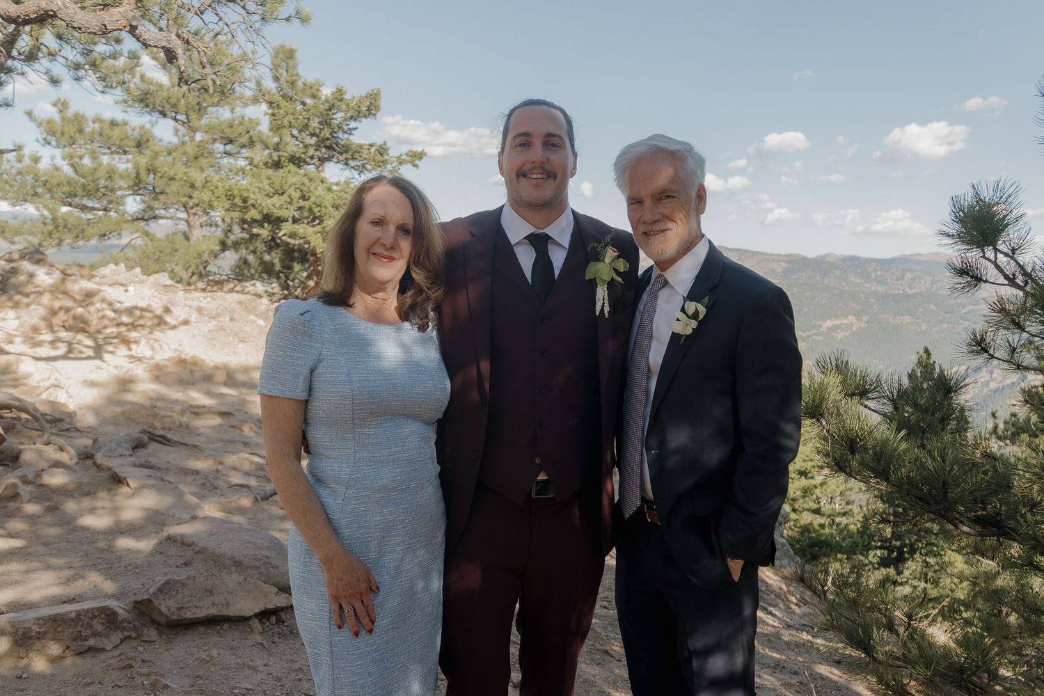 Groom in burgundy suit poses with two family members outdoors at a mountain wedding ceremony.