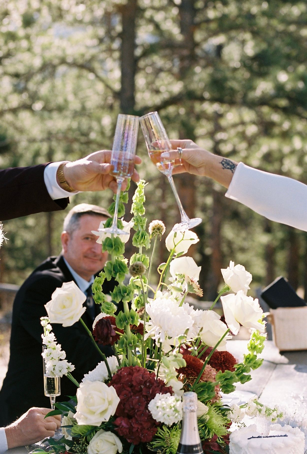 Wedding guests clink champagne glasses over a lush floral centerpiece of white and burgundy blooms outdoors.