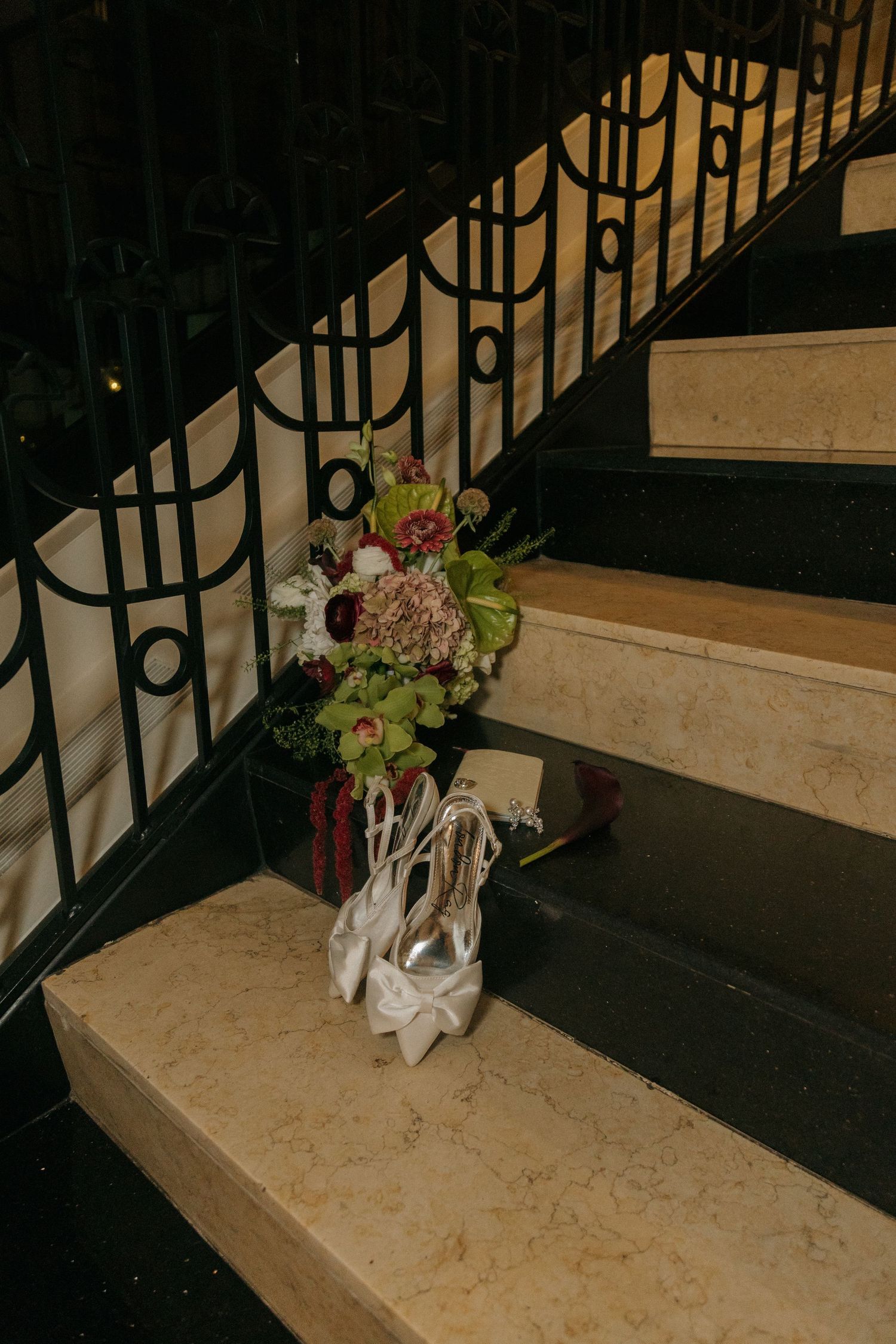 Bridal heels and floral bouquet with green orchids and burgundy blooms displayed on marble staircase with ornate black railing.