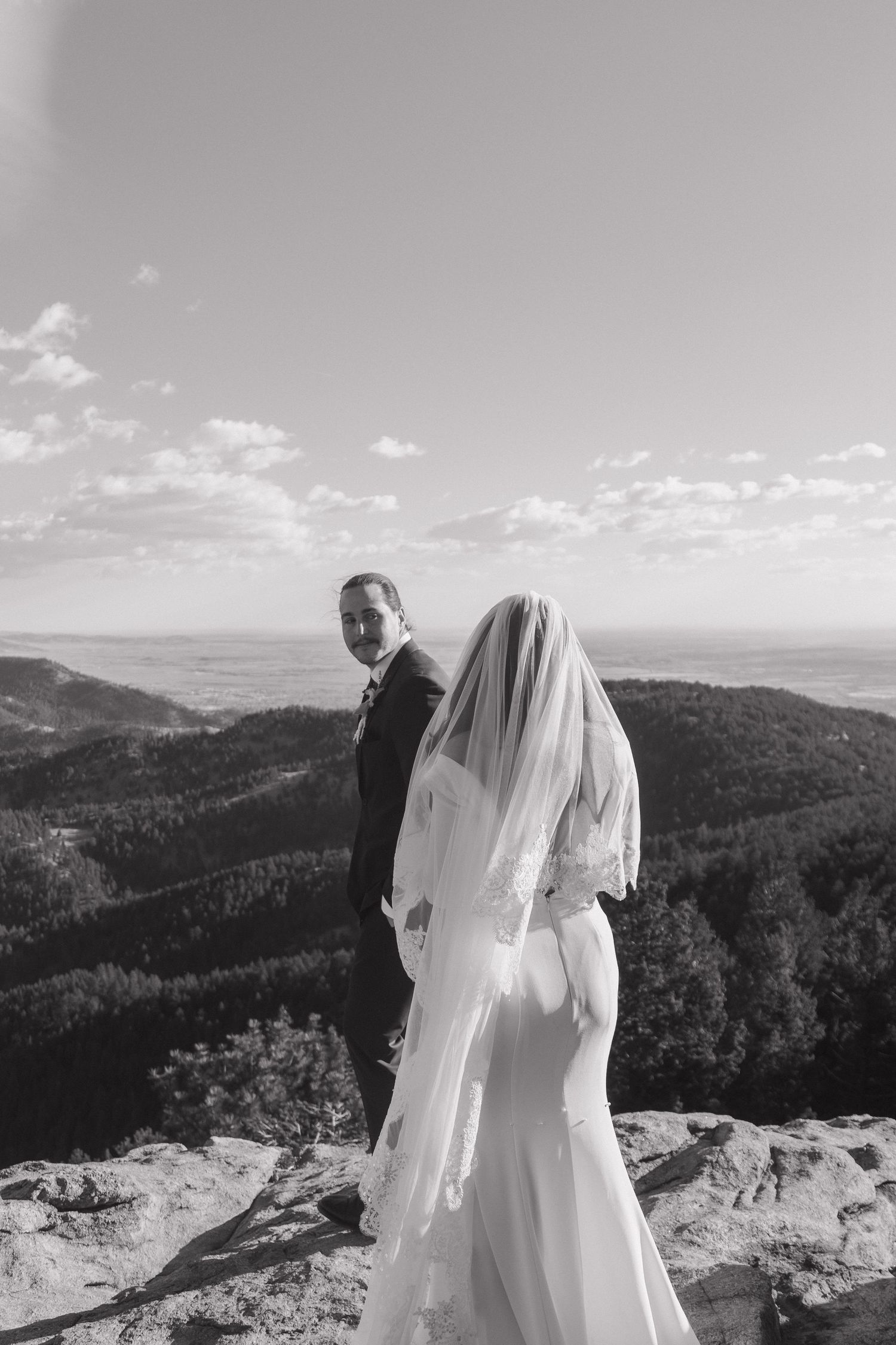 Bride and groom stand on mountain summit with sweeping valley views in black and white.