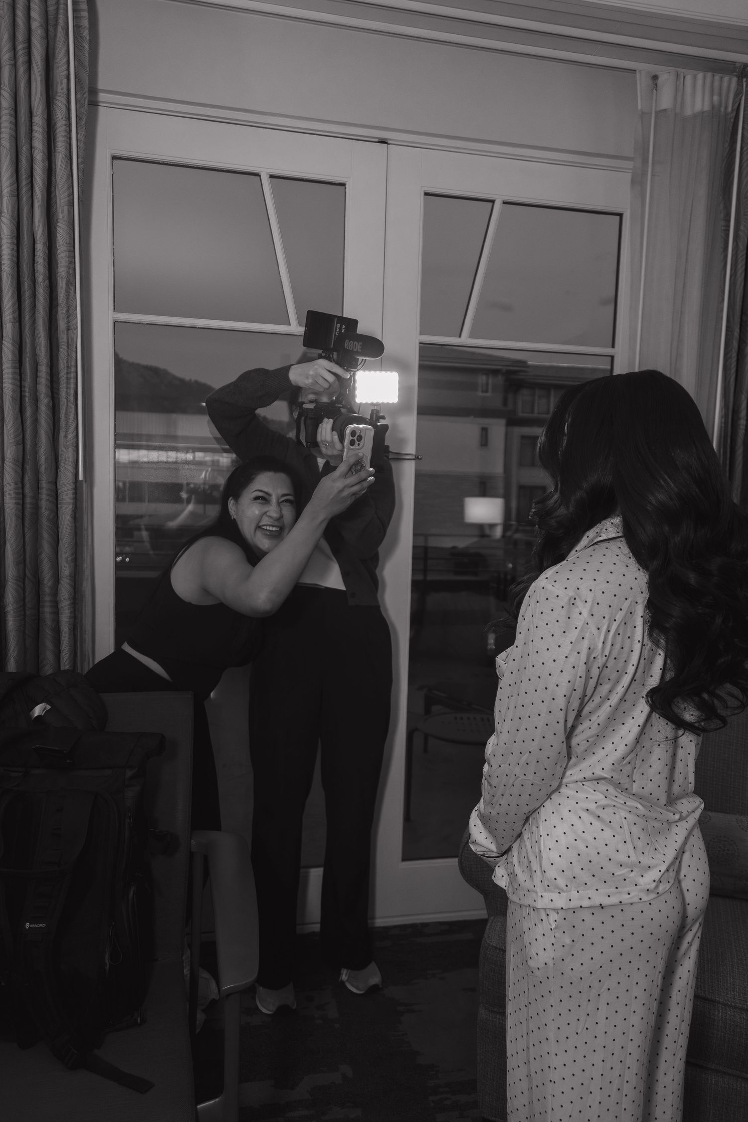 Black and white photo of a woman photographing a pregnant woman in a hotel room with a flash.