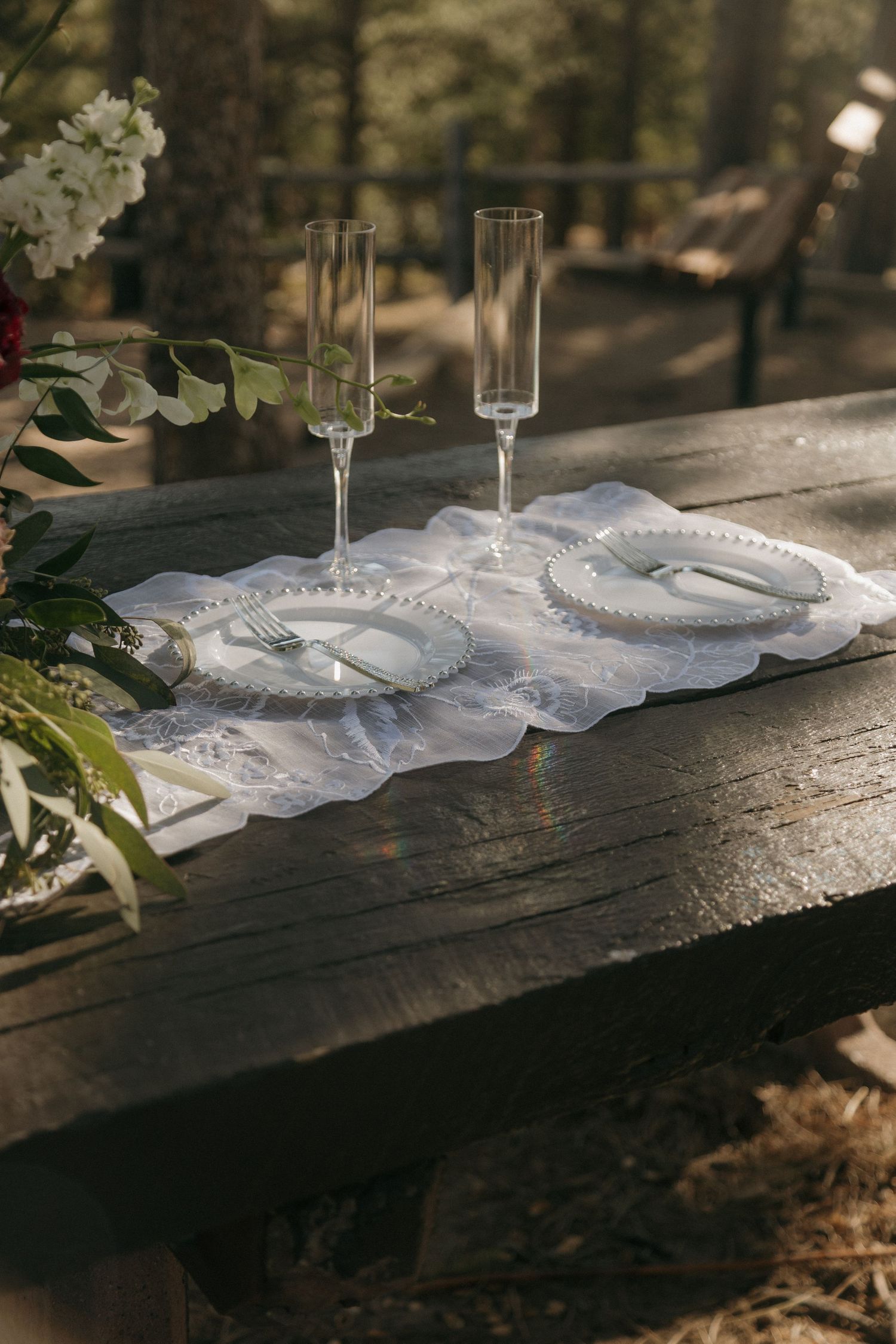 Rustic wooden table set for two with crystal champagne flutes, beaded plates, and white lace runner in forest setting.