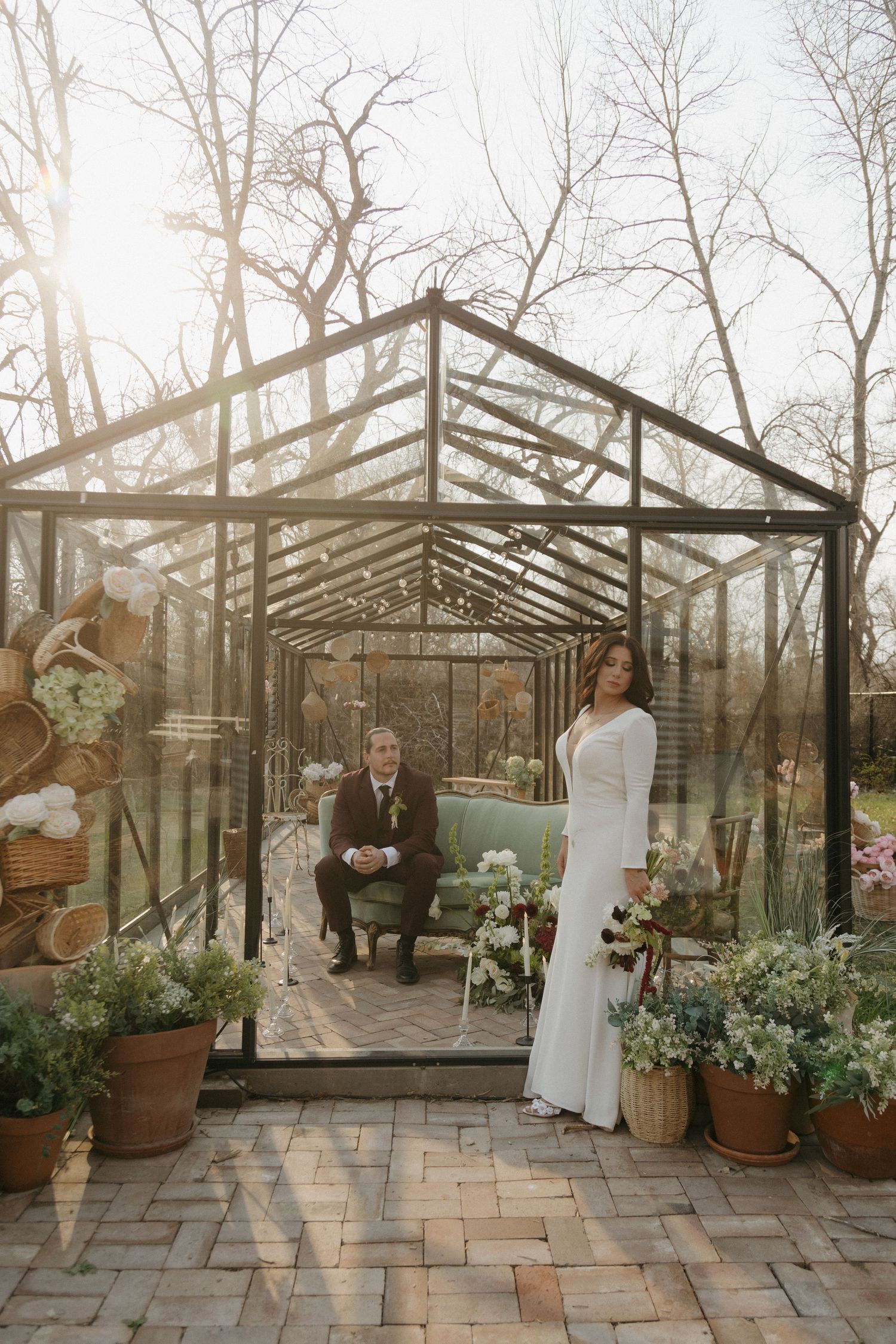 Bride and groom pose inside a sunlit glass greenhouse surrounded by potted plants.
