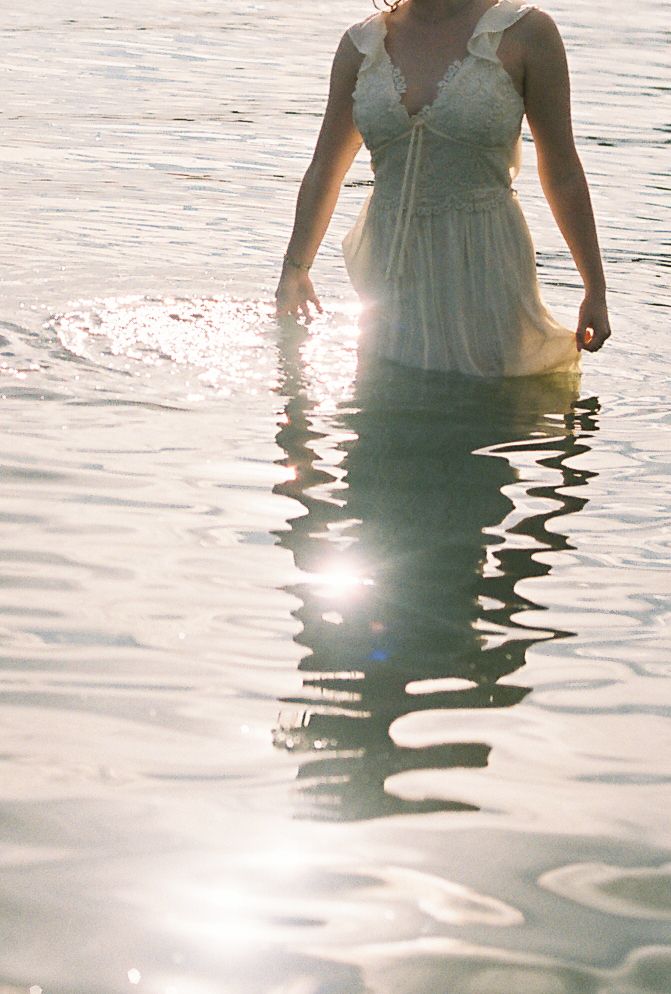 A bride in a white lace gown stands in shimmering sunlit water, her reflection visible below.