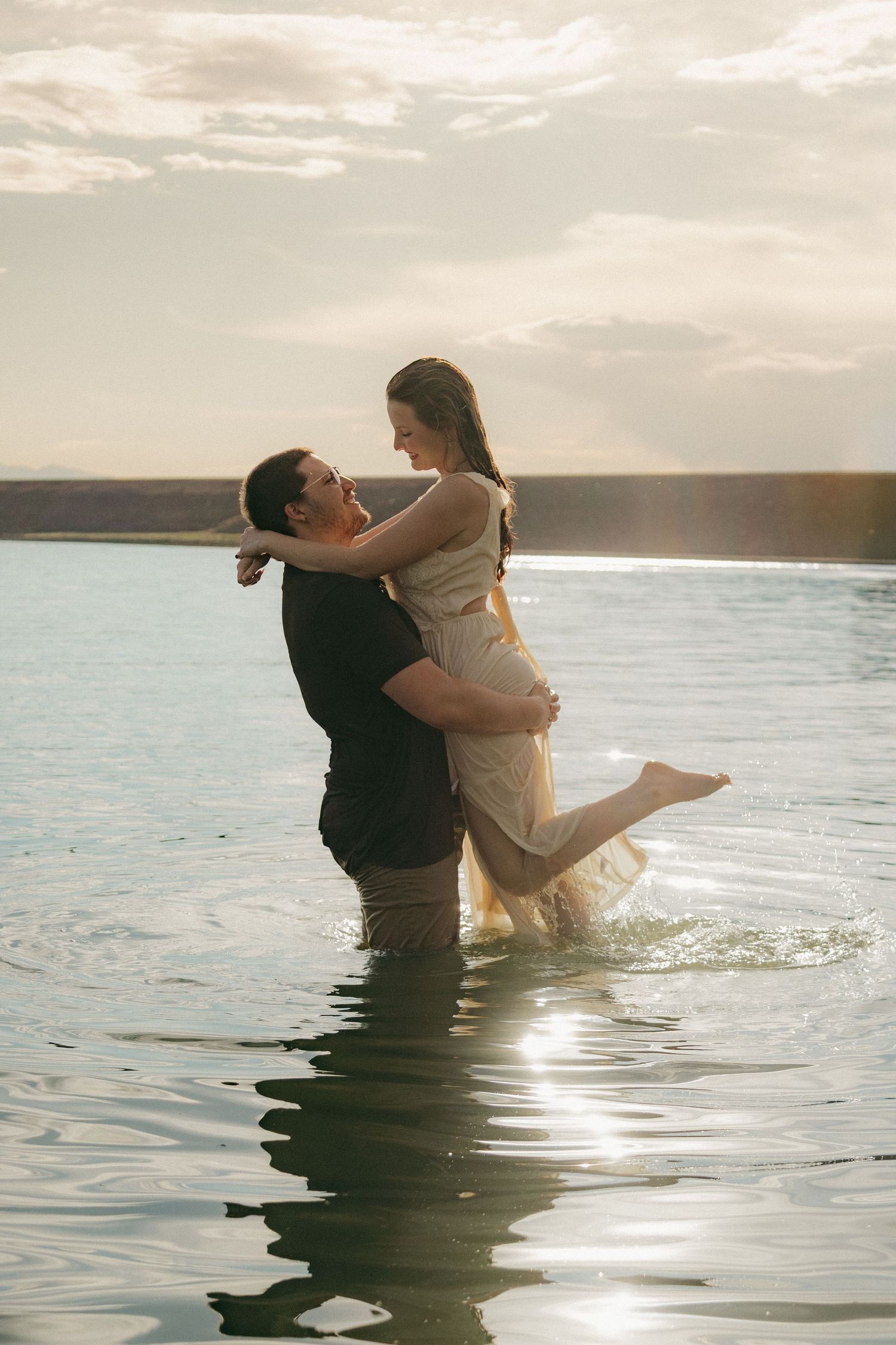Man lifting woman in white dress while standing in a lake with golden sunset light behind them.