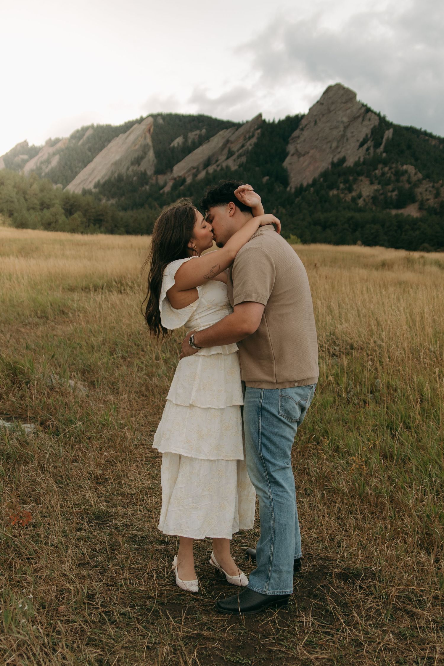 Couple shares romantic kiss in golden field with dramatic rocky Flatirons mountains in background.
