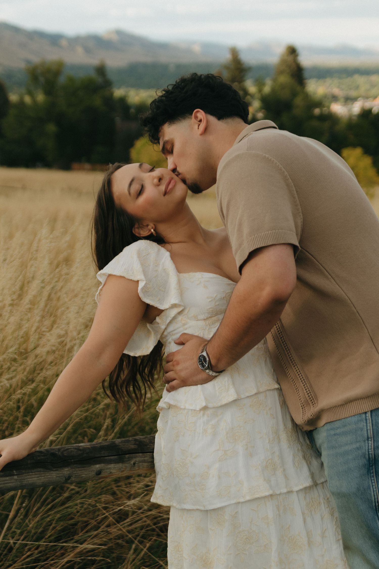 A couple sharing a romantic kiss in a golden field with mountain views during an outdoor engagement session.